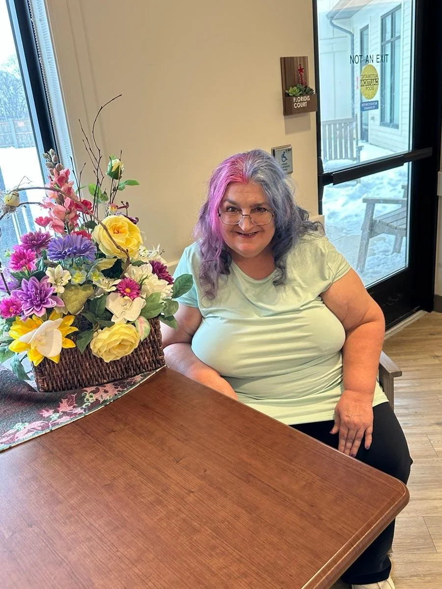 A woman with on gray and pink hair and glasses sitting at a table with a basket of colorful flowers, smiling at the camera. Behind her is a glass door with signs, one indicating 'Not an Exit' and a view of snow outside.