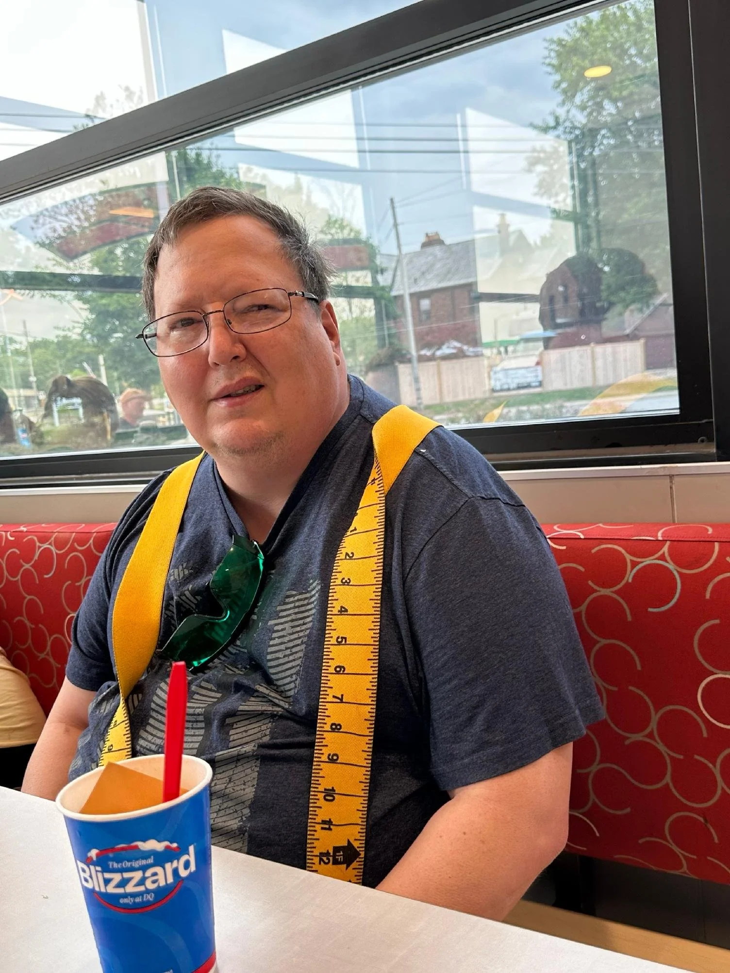 Man sitting inside a restaurant, wearing glasses and a dark gray T-shirt, with a yellow measuring tape draped over his shoulders. There is a blue soda cup with a red straw on the table. The background shows a window with outdoor trees and houses.