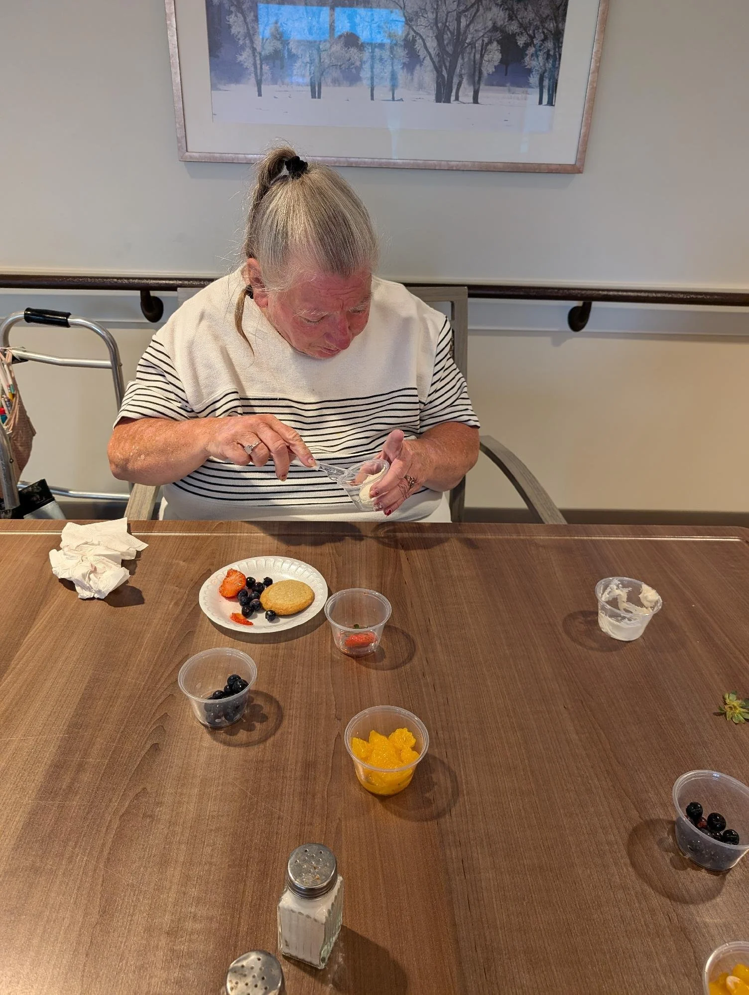 An elderly woman with blonde hair tied in a ponytail, wearing a striped shirt, is sitting at a wooden table with small bowls of fruit, including blueberries and orange slices, and a plate with a cookie and berries. She is scooping food from a small c