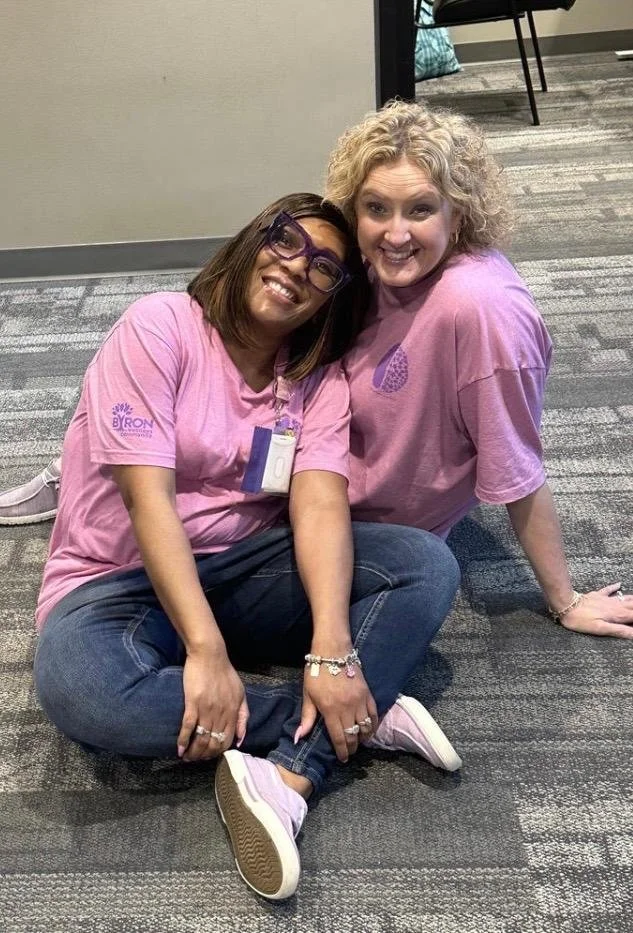 Two women sitting on the floor, smiling at the camera. One woman has dark hair, glasses, and is wearing a pink T-shirt, while the other woman has curly blonde hair and is wearing a pink T-shirt with a purple design. They are in an indoor setting with