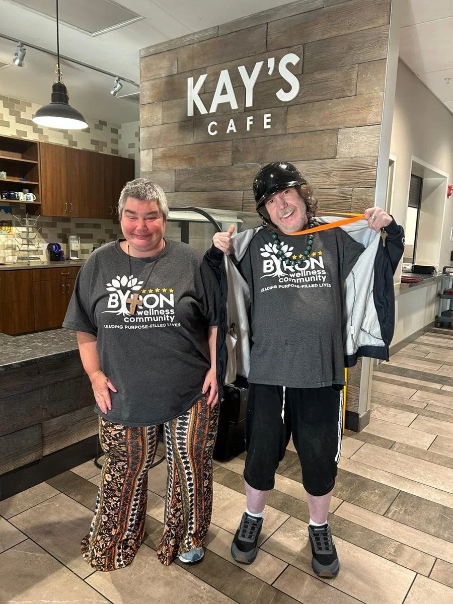 Two people standing inside a cafe, smiling. The woman on the left is wearing a gray t-shirt with the words "BYON wellness community". The man on the right is wearing a helmet, gray t-shirt with the same logo, and a jacket draped over his shoulders, h