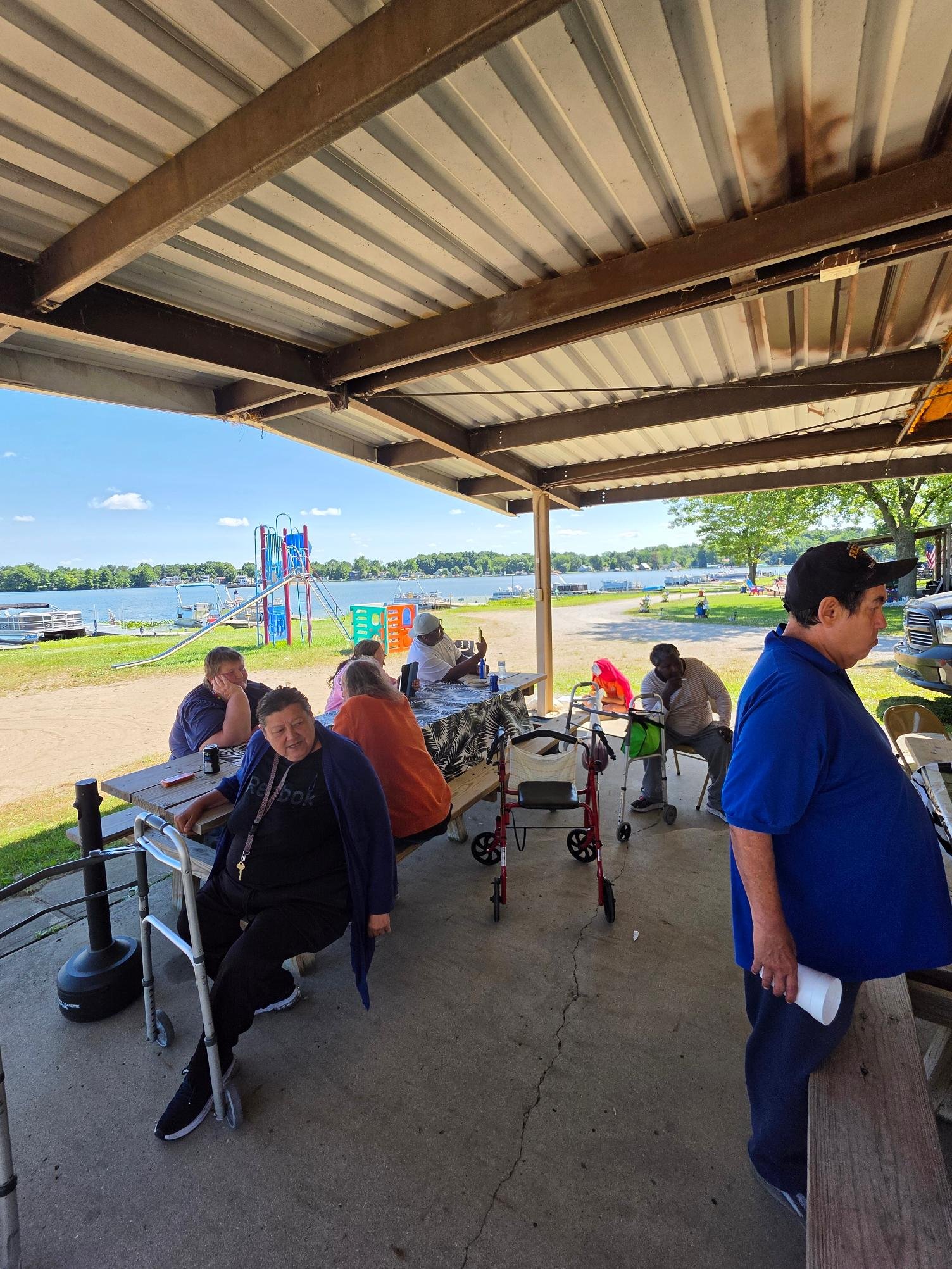 People gathered under a pavilion near a lake, with a slide and play area in the background. Some are sitting at picnic tables, others are standing or using phones, with trees and boats visible across the water.