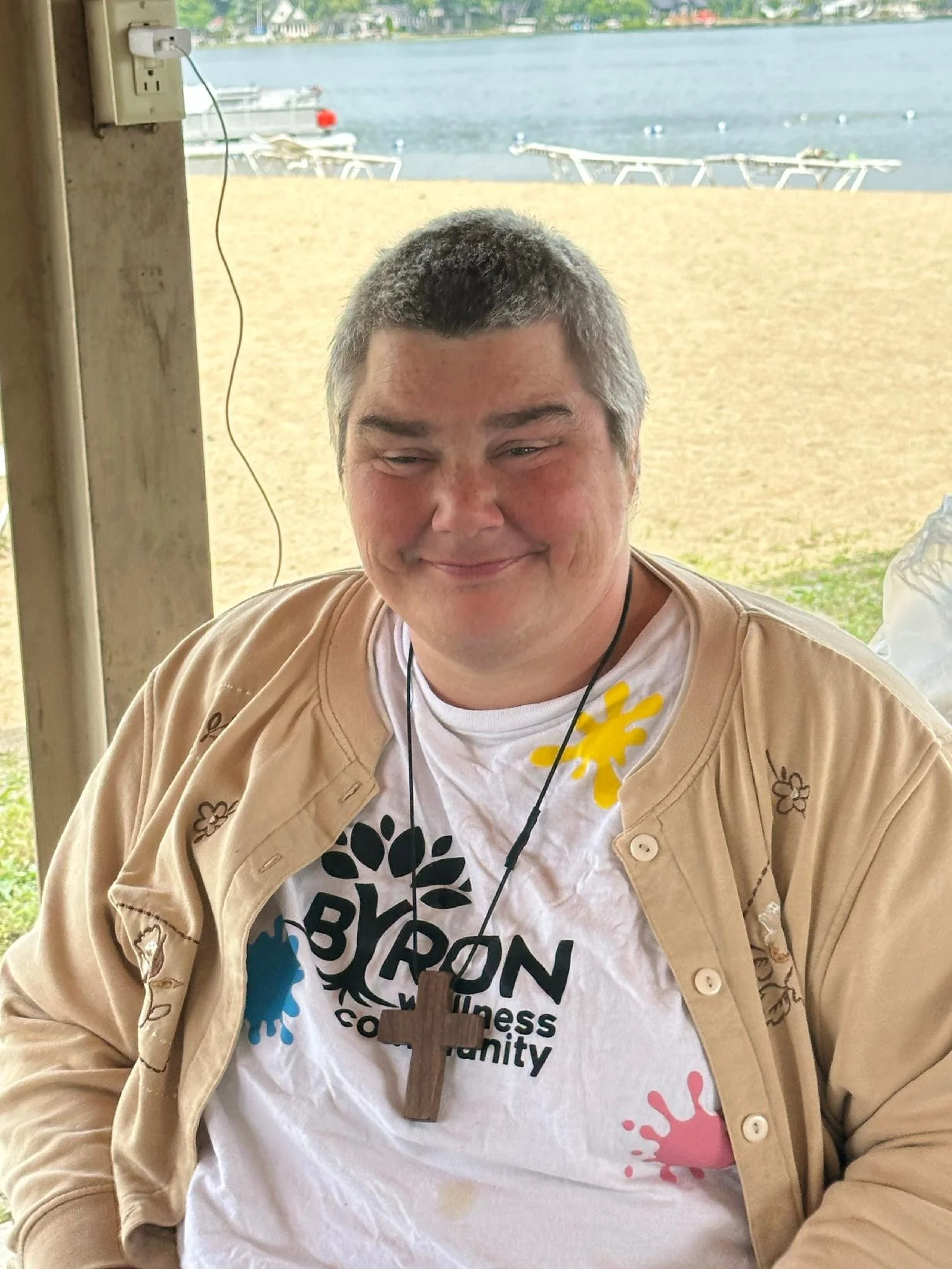 A person wearing a white T-shirt with a logo and text, a tan button-up shirt, and a wooden cross necklace, sitting near a window with a lakeside view and boats in the background.