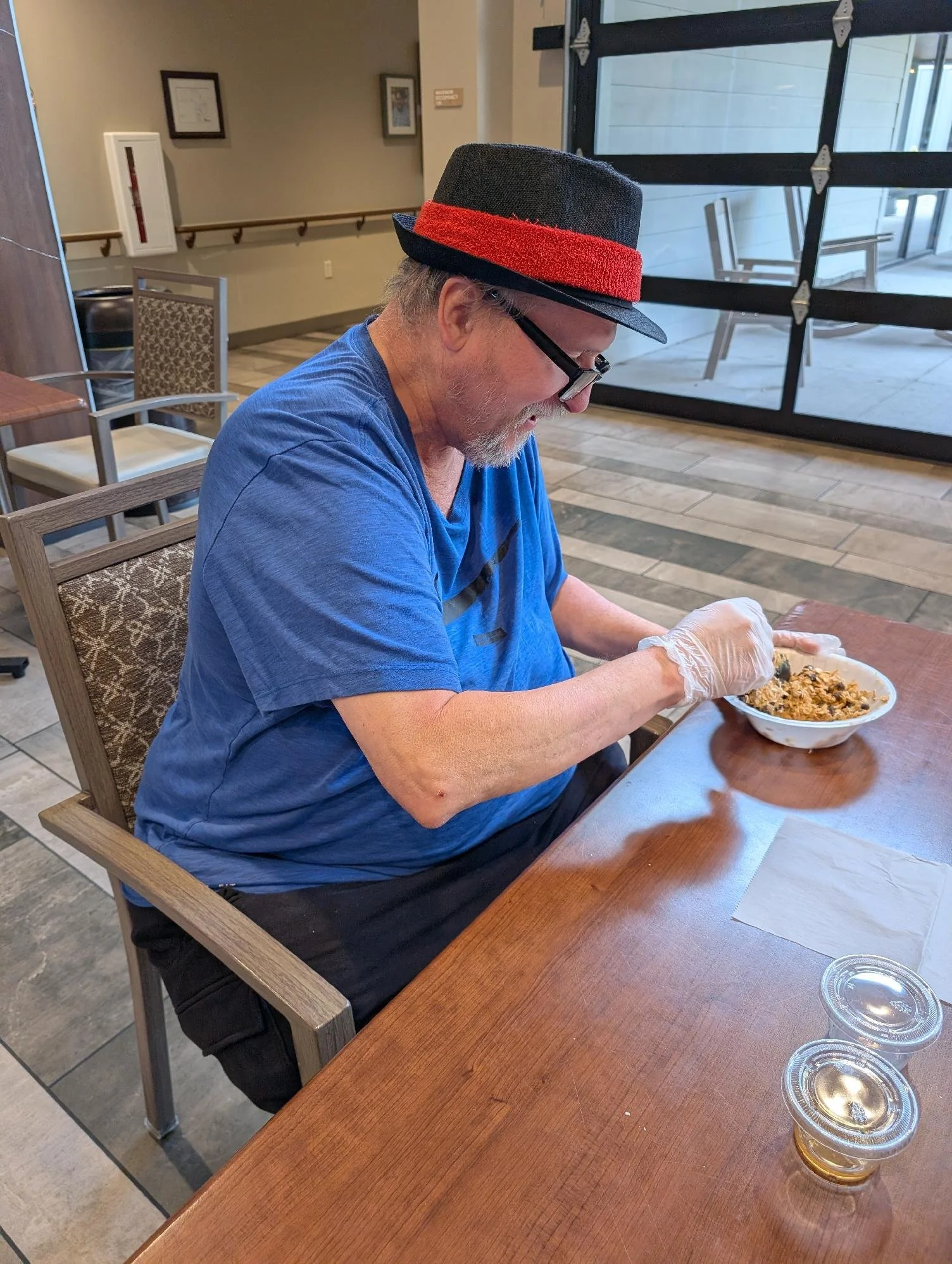 An elderly man wearing a black hat with a red band, glasses, and a blue shirt, sitting at a wooden table, eating food from a bowl with gloves on his hands, in a restaurant setting.