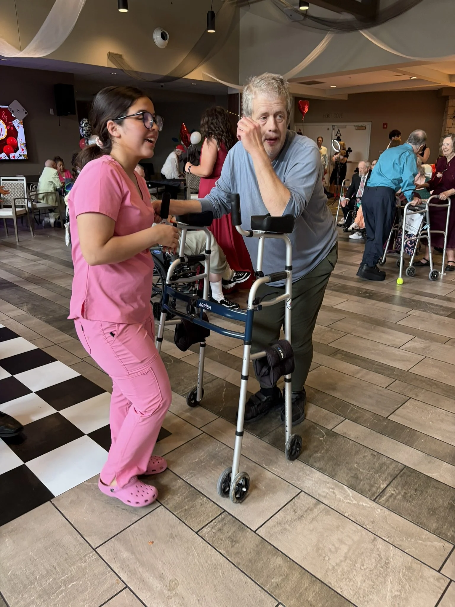 A young woman wearing pink scrubs and glasses is smiling and talking to an older man with gray hair and a beard, who is leaning on a walker. The scene appears to be at a party or social gathering with other elderly people in the background, some usin