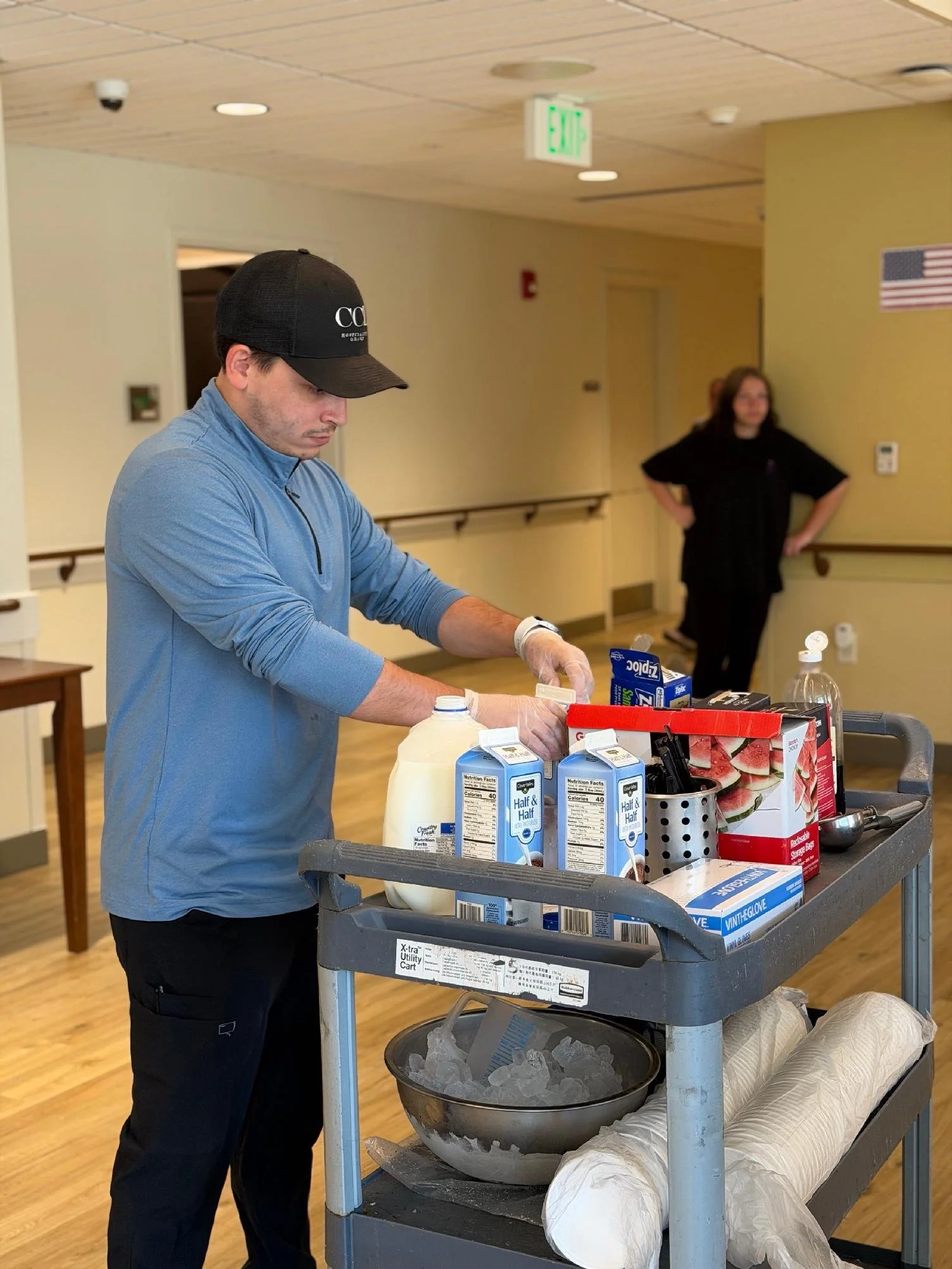A man in a blue jacket and black cap working at a food service cart filled with dairy products, paper towels, and food boxes. In the background, a woman in black stands with her hands on her hips in a room with yellow walls, American flag, and exit s