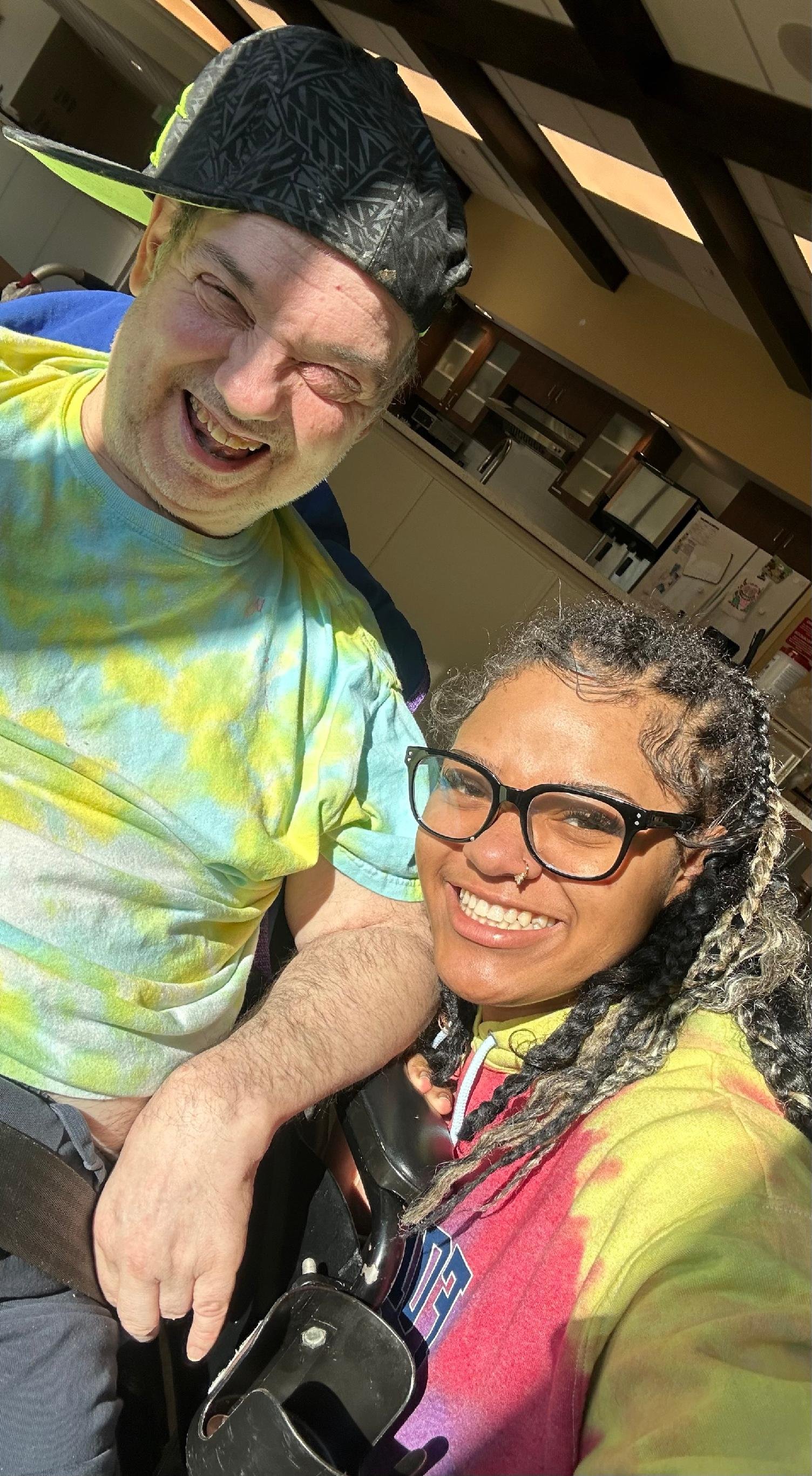 Two people smiling and taking a selfie in a kitchen, with one wearing a black cap and colorful tie-dye shirt, and the other with glasses, braided hair, and a red and yellow tie-dye shirt.