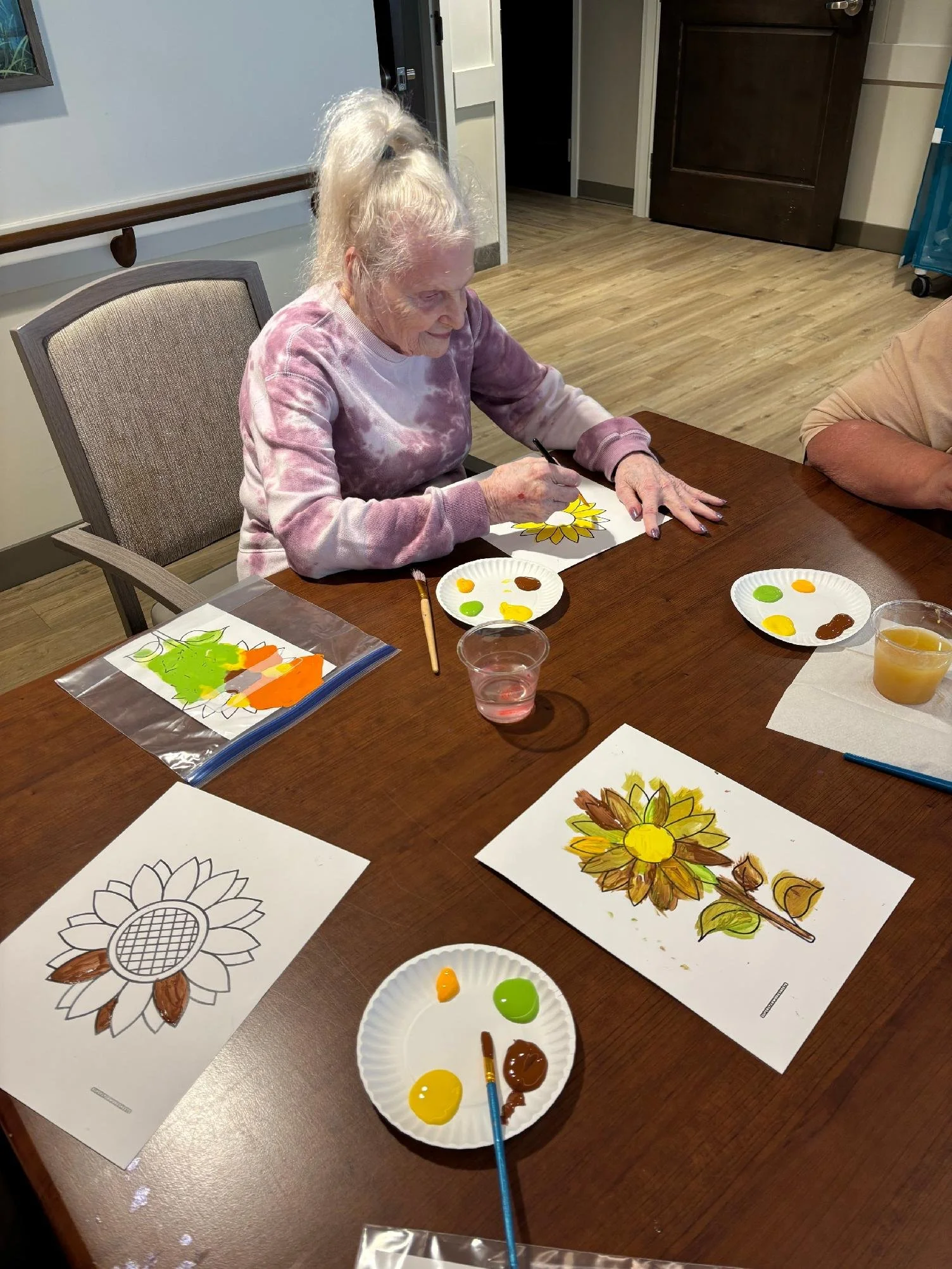 An elderly woman with white hair tied up in a high ponytail is sitting at a wooden table, painting a sunflower illustration with brown and yellow paint. Several paint palettes and coloring sheets are spread on the table, and she is happily focused on