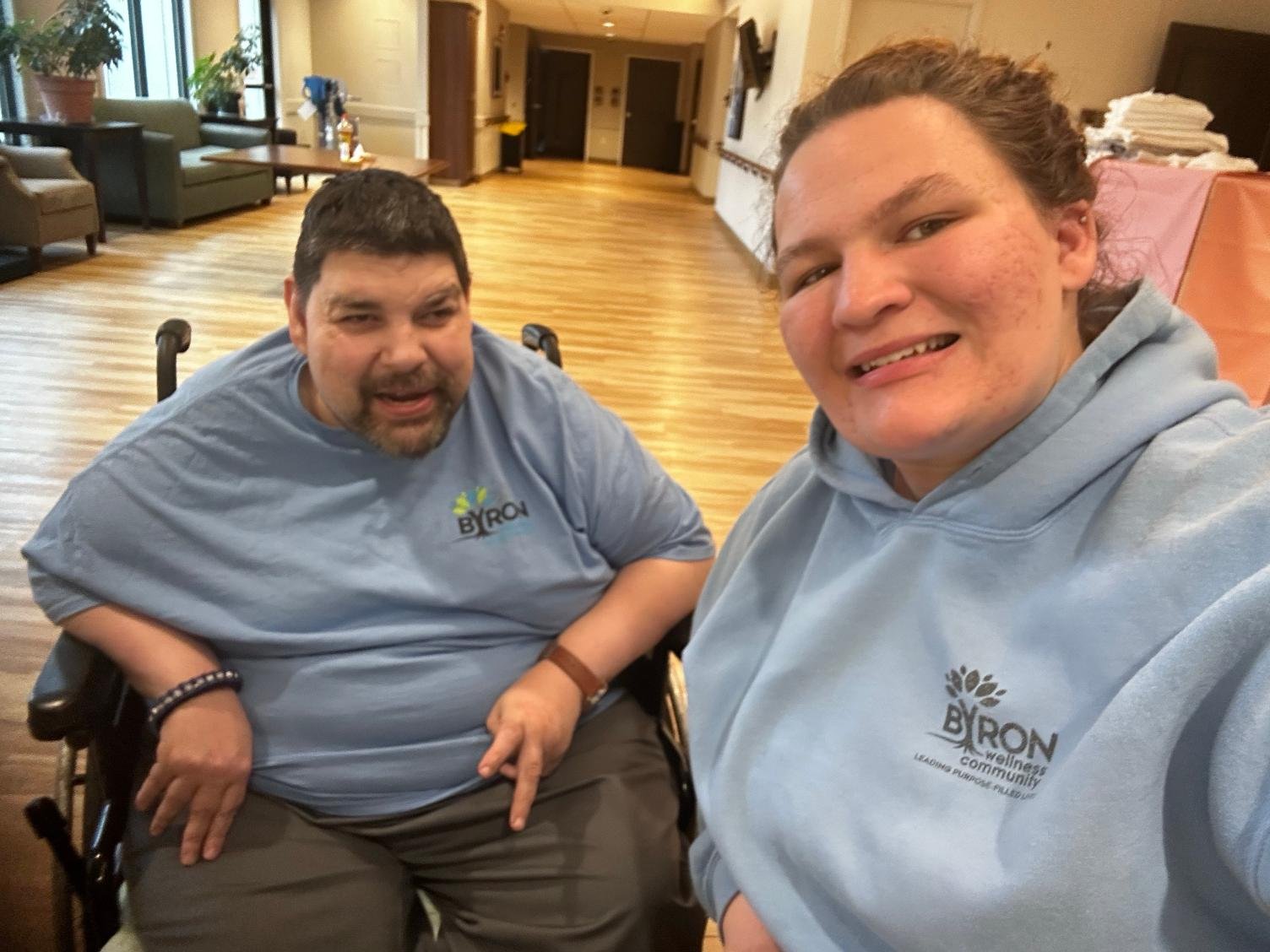 Two people taking a selfie indoors, both wearing light blue hoodies with a logo that reads 'Byron Wellness Community'. They are smiling and appear to be in a community or healthcare setting with wooden floors and comfortable seating in the background