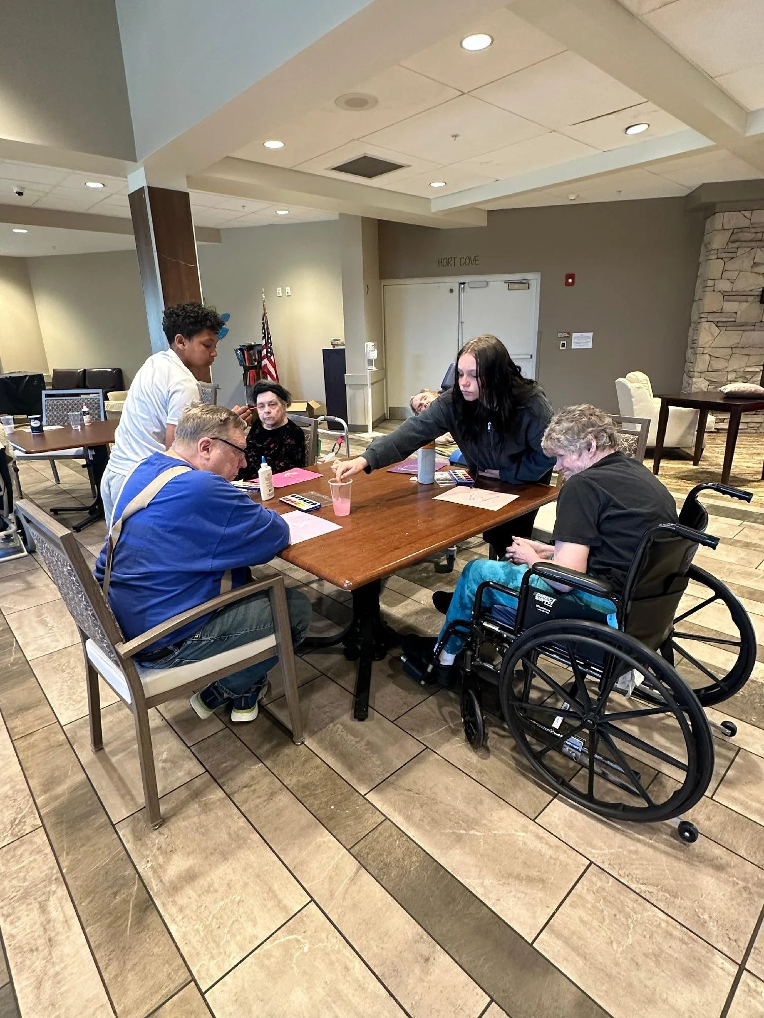 Group of seven people, including two in wheelchairs, gathered around a table in a room with beige tiled floors, a stone fireplace, and light green walls. Some appear to be engaging with each other, one woman is reaching across the table, and others a