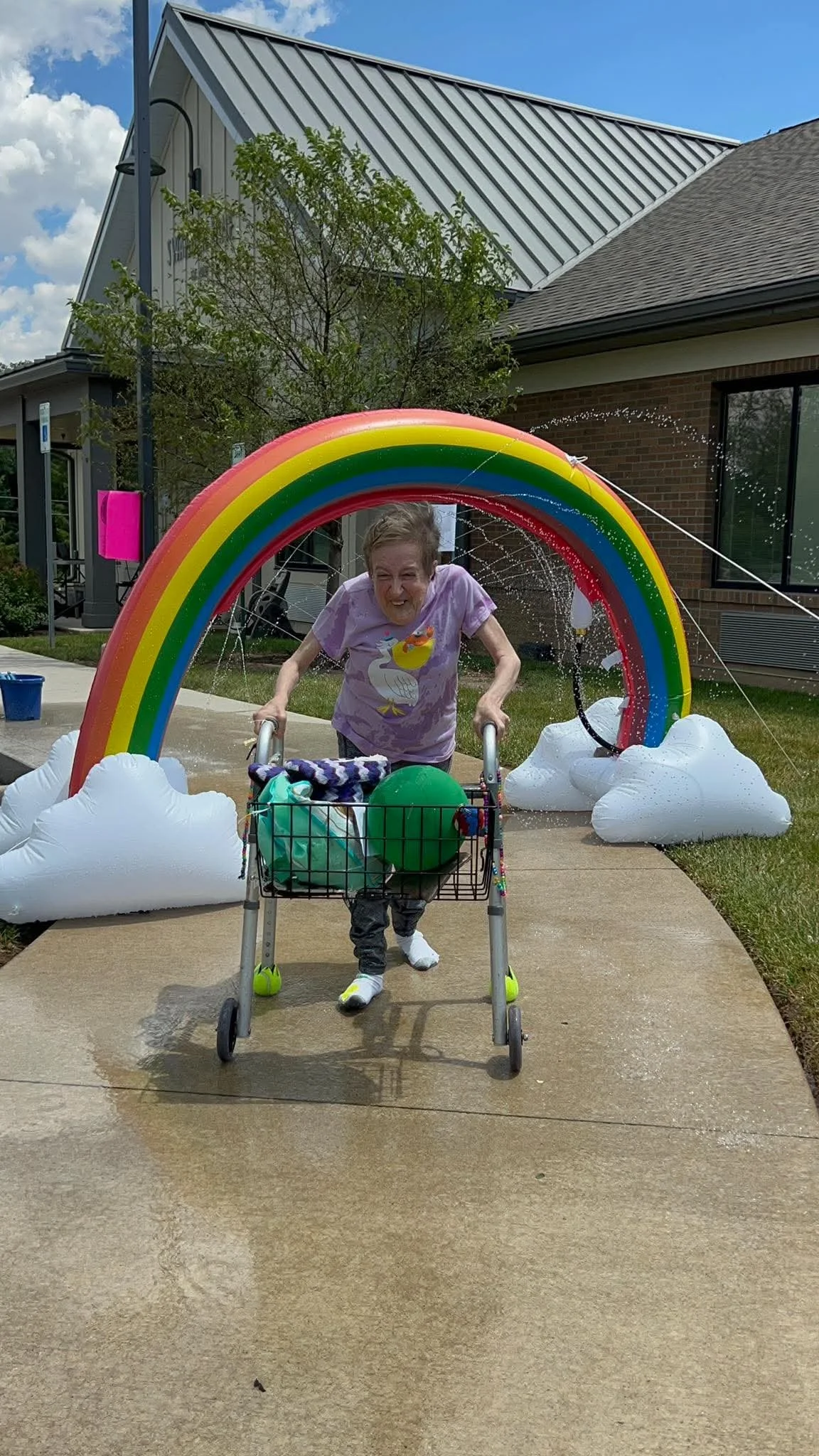 An elderly woman smiling and pushing a walker through a decorative archway shaped like a rainbow with clouds, outside a building, on a sunny day.