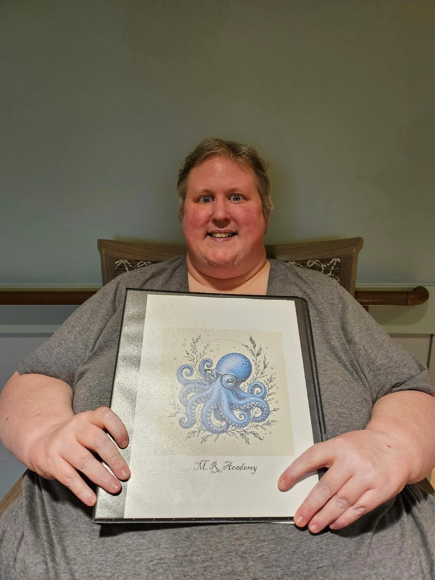 A smiling man with gray hair and a beard holding a framed artwork of a blue octopus with floral decorations, labeled 'M.R. Academy,' sitting at a table in front of a green wall.