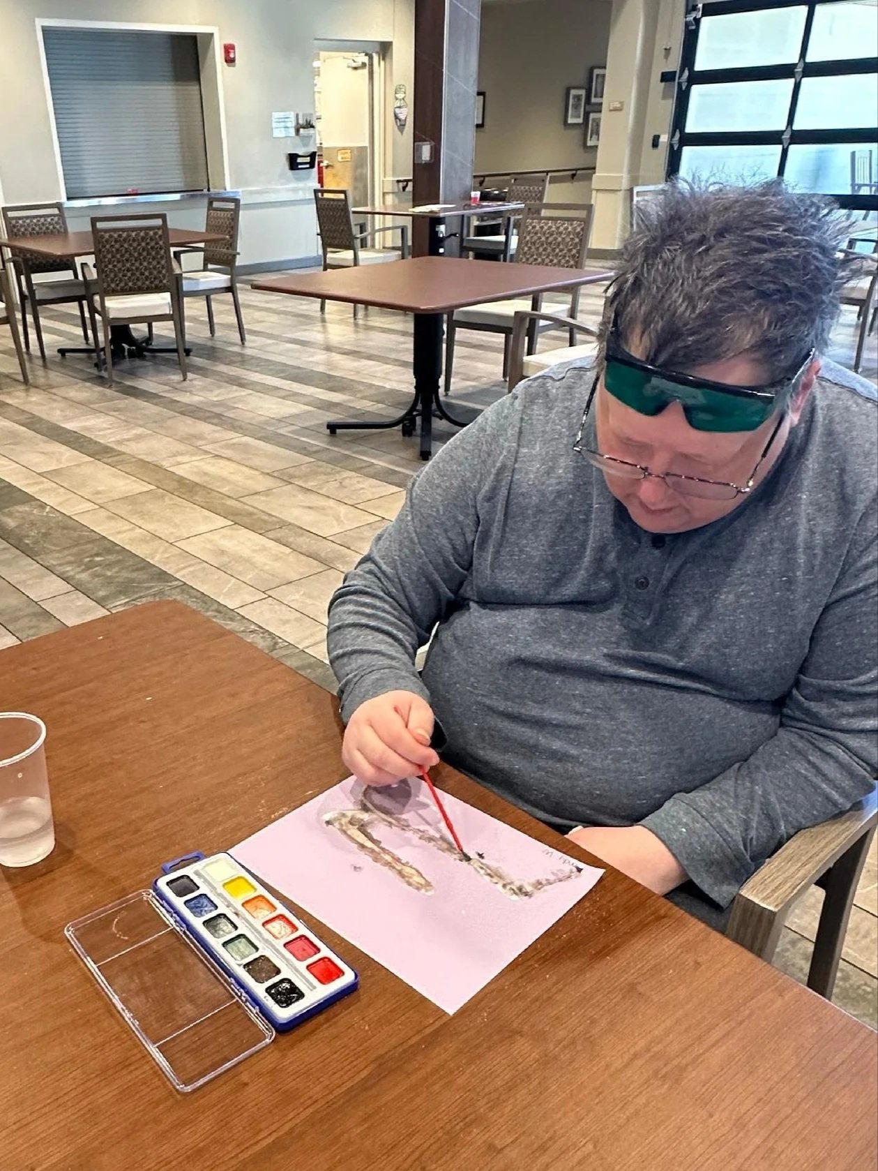 A man wearing glasses and a gray long-sleeve shirt sitting at a wooden table, painting with watercolors on pink paper in a cafe or restaurant with empty tables and chairs in the background.