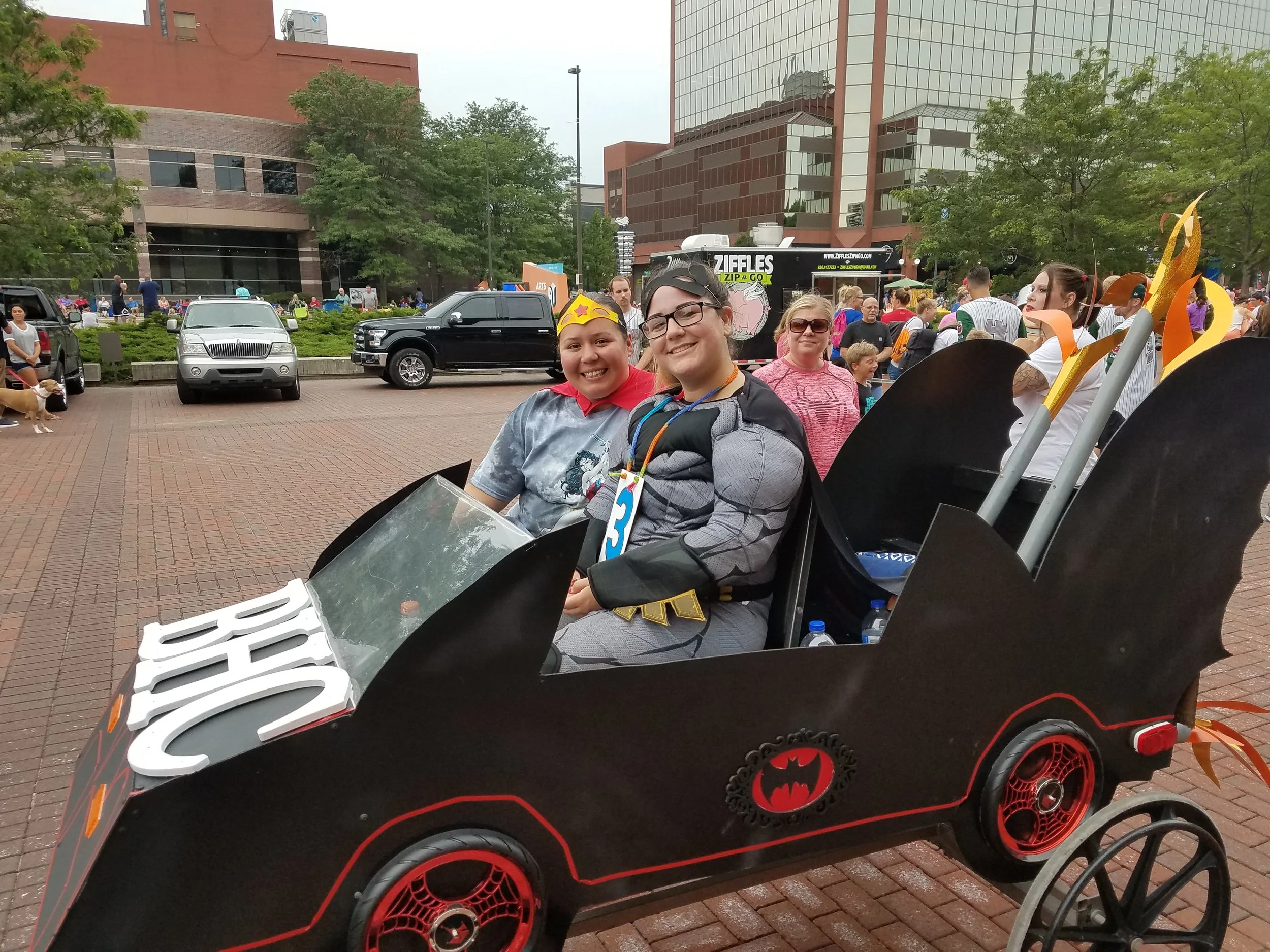 Three smiling people sitting on a Batman-themed ride at an outdoor event or fair, surrounded by trees, cars, and a food truck in the background.