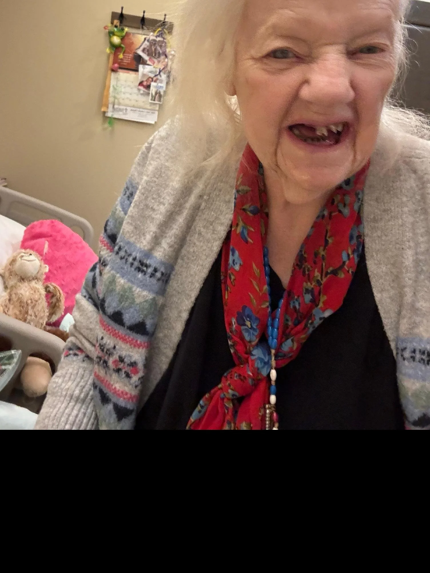 A close-up of an elderly woman with gray hair, wearing a red scarf with a floral pattern and a gray sweater with traditional patterns. She appears to be smiling or talking, with visible teeth.