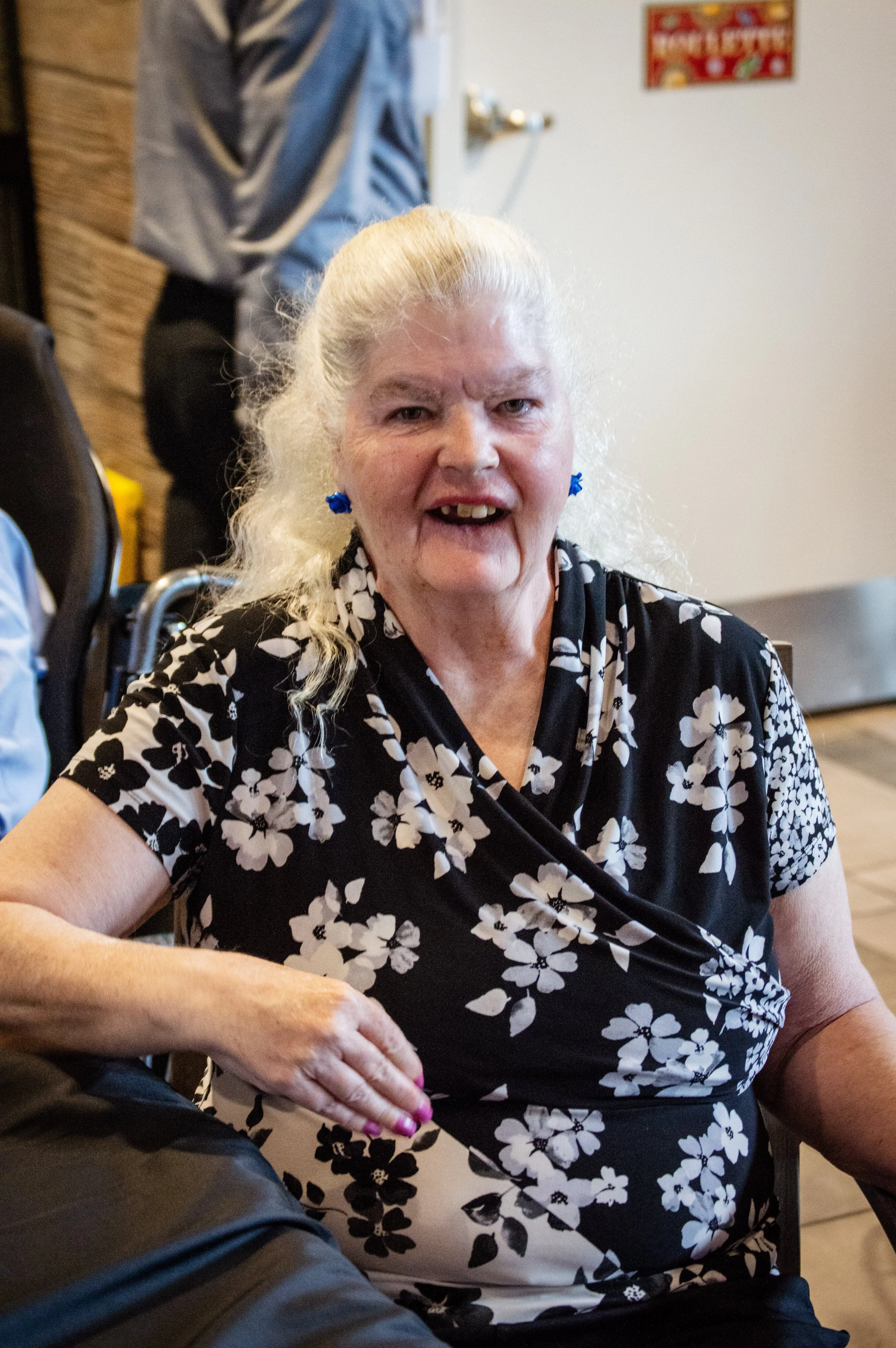 A smiling elderly woman with long white hair tied back, wearing a black and white floral patterned blouse and blue earrings, sitting at a table.