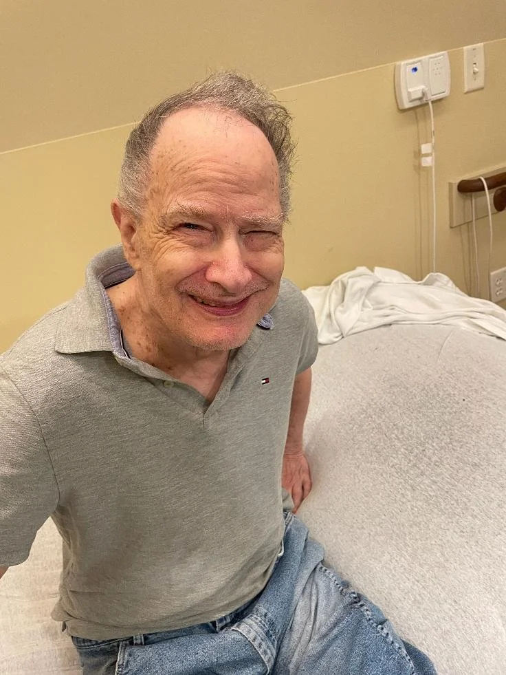An elderly man with a big smile, sitting on a hospital bed in a room with beige walls, wearing a gray polo shirt and jeans.