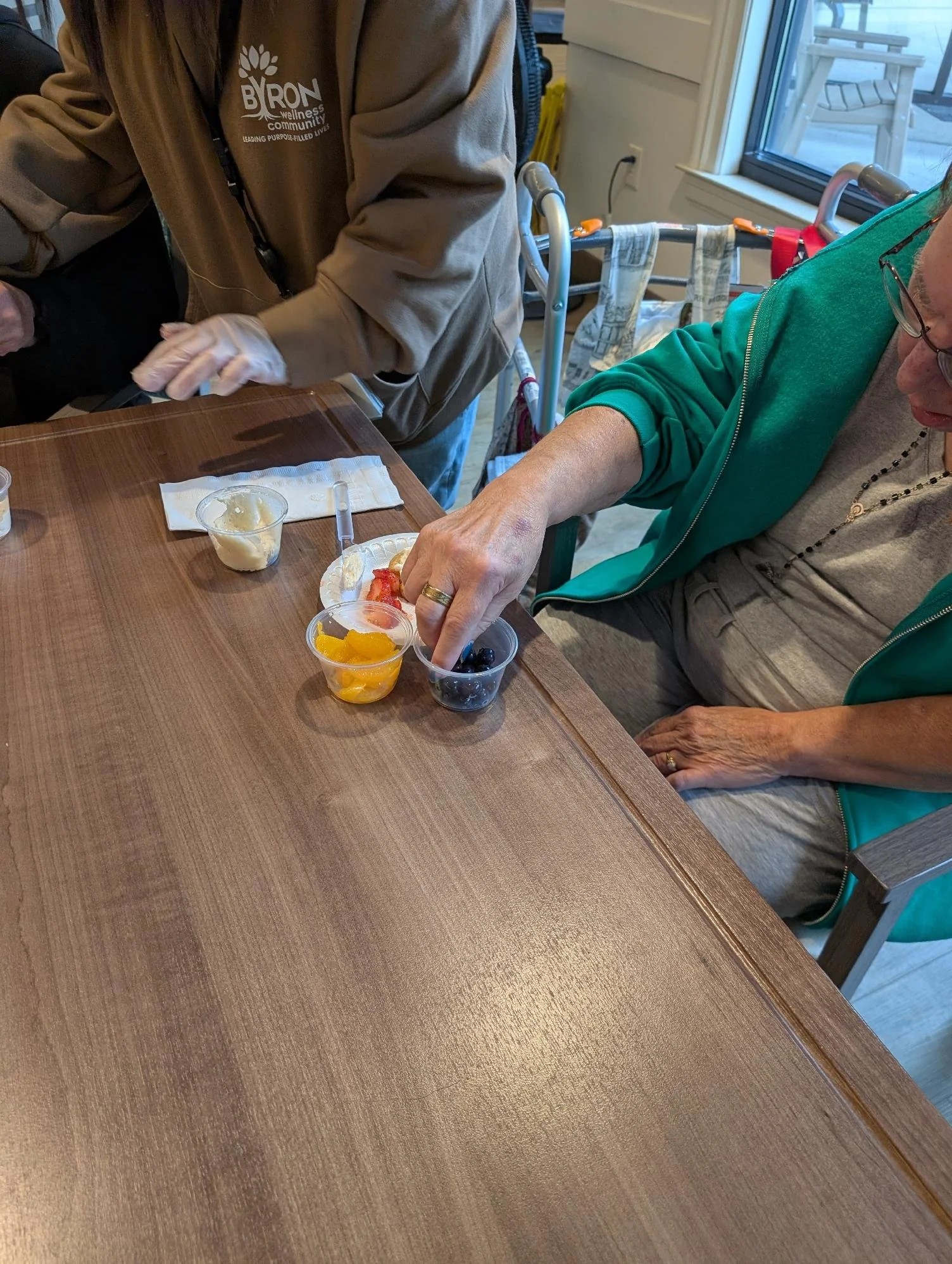 A person with gray hair and glasses is reaching for a small plastic cup of fruit on a wooden table. Another person, wearing a beige jacket, is standing nearby. A window with a sliding door, a walker, and a towel hanging on it are visible in the backg
