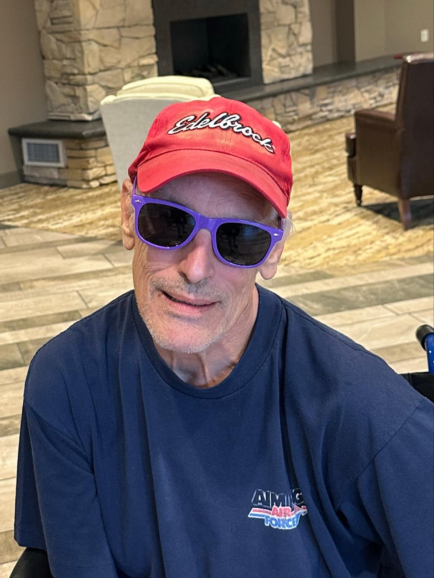 A man wearing a red cap with the word 'Edelbrock' and purple sunglasses, sitting indoors near a stone fireplace, smiling and looking slightly to the left.