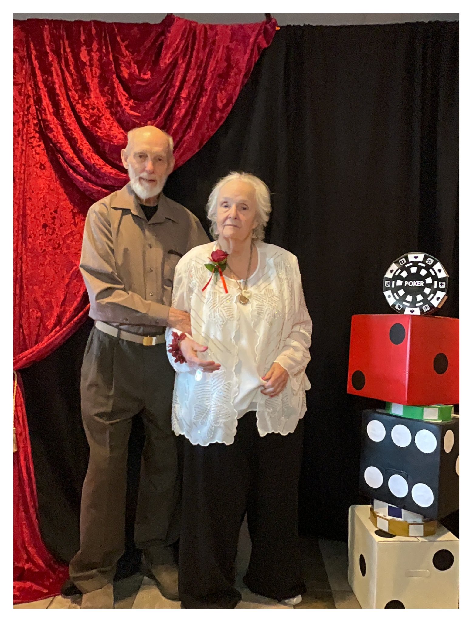 An elderly man and woman posing together in front of a red and black backdrop, with large dice props beside them. The woman is holding a small object, possibly a cake or a gift, and is wearing a white dress with a red corsage. The man is wearing a br