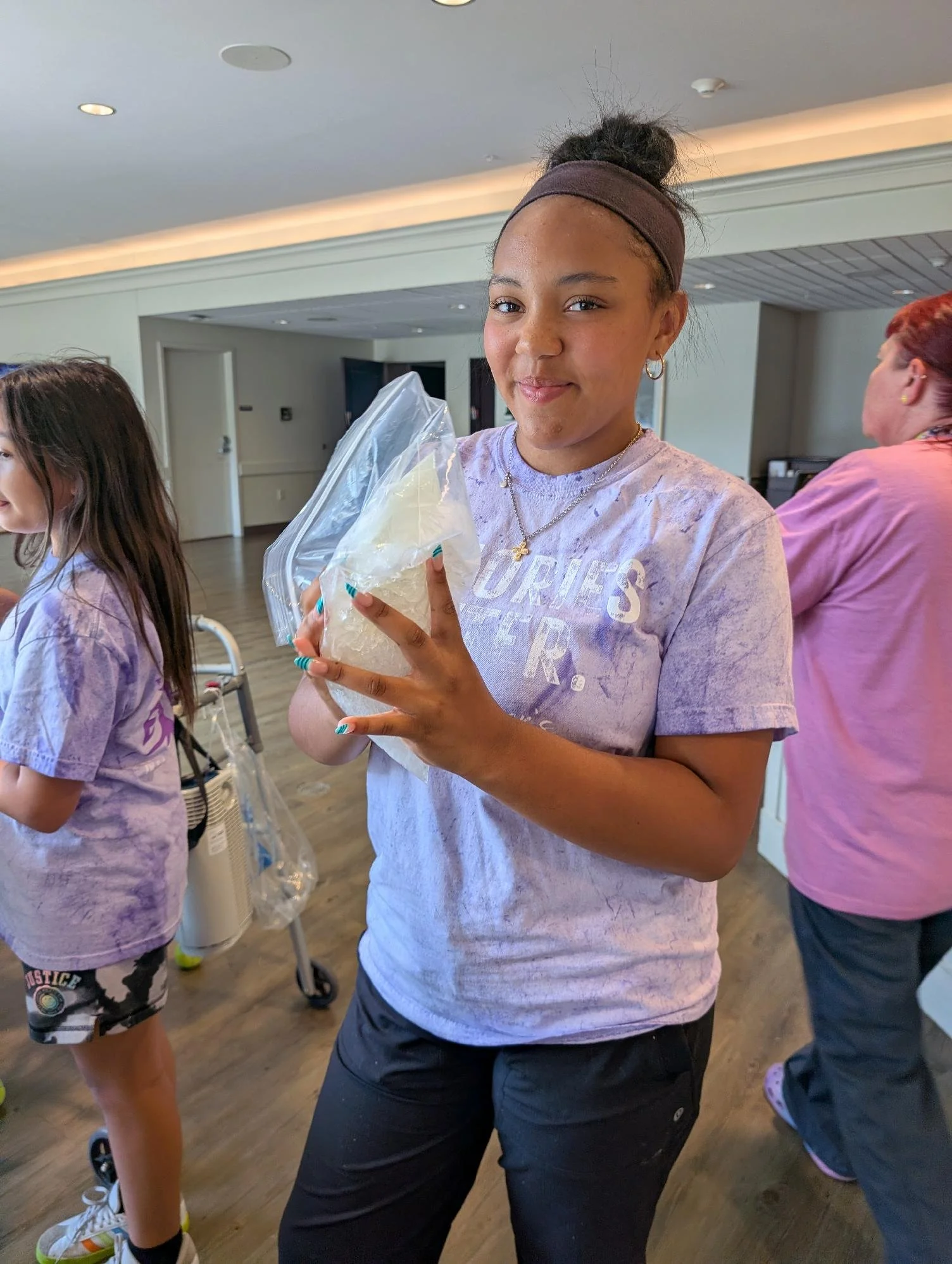 A young girl with curly hair tied in a bun, wearing a headband, jewelry, a tie-dye t-shirt, and black pants, holding a plastic bag with a yellow object inside, standing indoors with other people around.