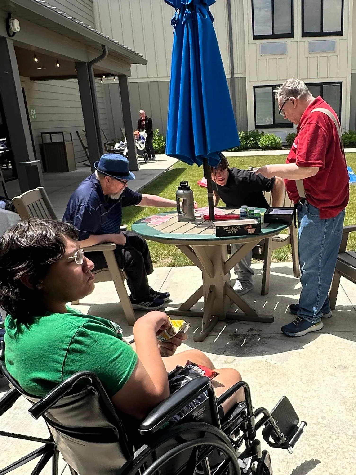 People at an outdoor gathering, one in a wheelchair, others sitting at a table with an umbrella, engaging in activities like reading and talking.