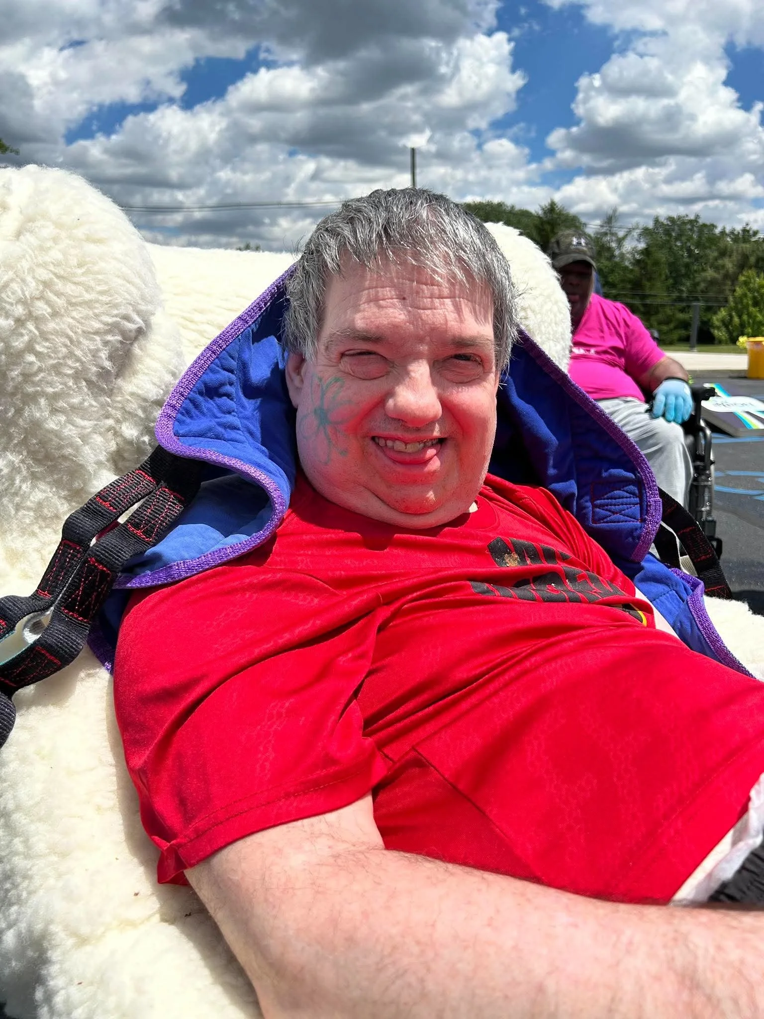 Close-up of a smiling man with face paint, lying on a soft surface, with another person in a wheelchair and a blue sky with clouds in the background.