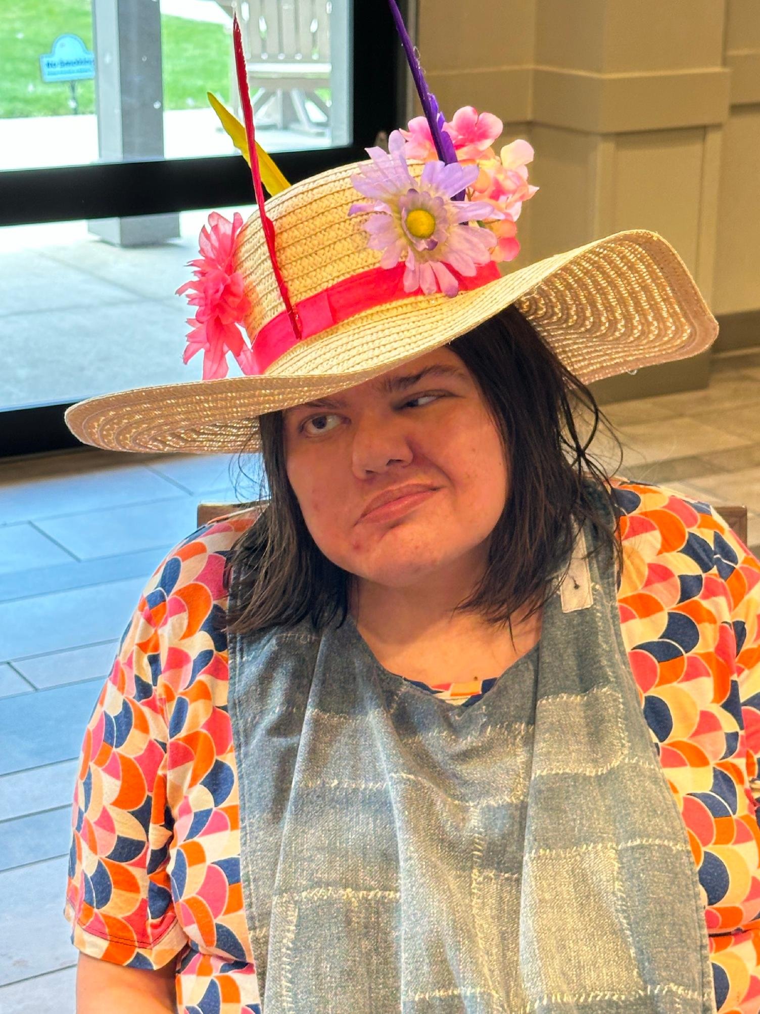 A person wearing a wide-brimmed straw hat decorated with pink and purple artificial flowers, red and yellow feathers, and a red ribbon. They are inside near a window, wearing a colorful outfit with a patterned top and a metallic apron.
