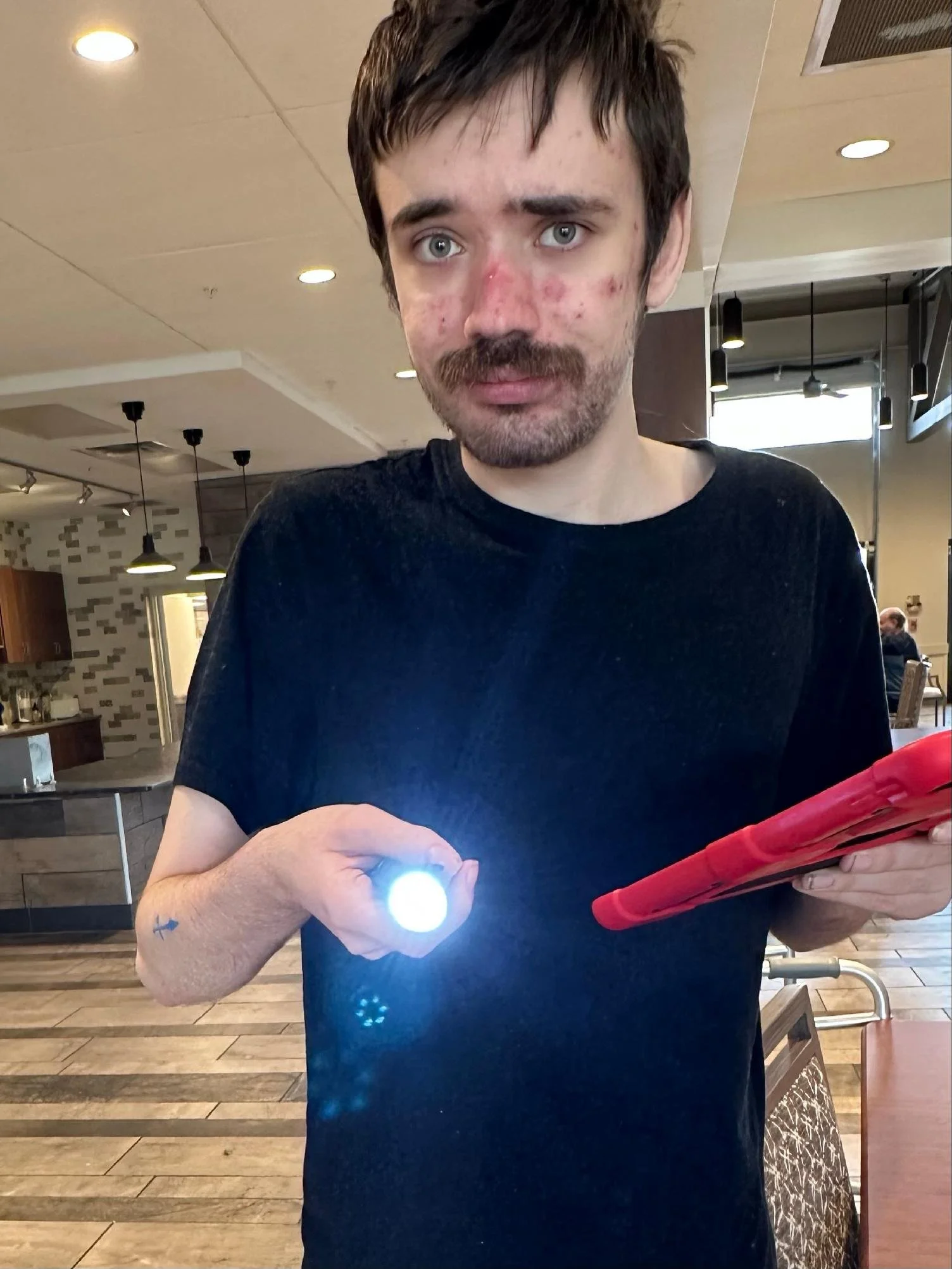 Young man with short dark hair, mustache, and acne on face, holding a flashlight in his right hand and a red tablet in his left hand, standing inside a restaurant or cafe.