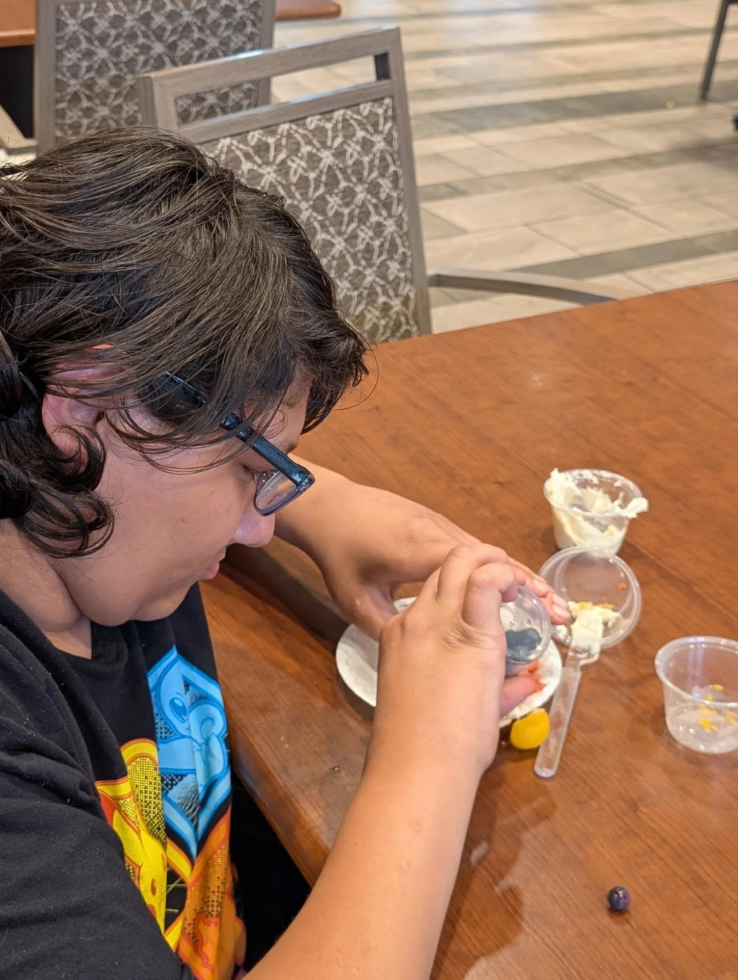 A person with curly brown hair, wearing glasses and a black shirt with colorful design, is sitting at a wooden table in a restaurant, eating ice cream from a clear plastic cup.