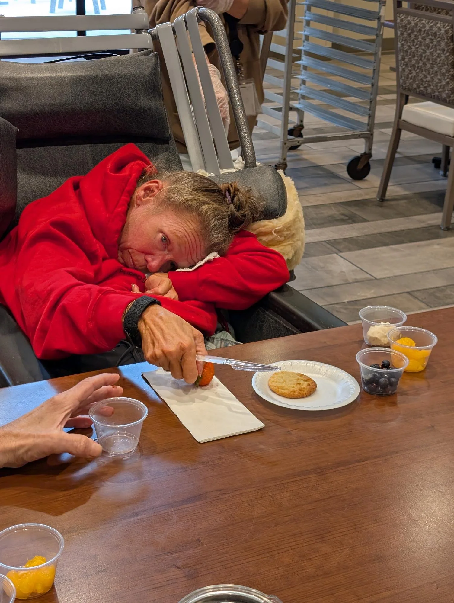 An elderly woman wearing a red hoodie is lying on a bench, resting her head on a cushion while eating strawberries at a table with various desserts, including cookies, fruit cups, and whipped toppings.