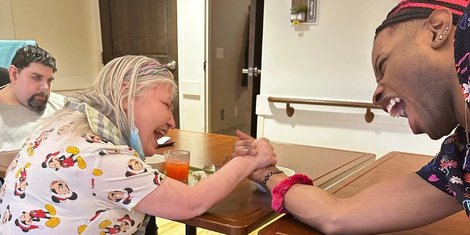 Two women are engaging in an arm wrestling match at a table, smiling and laughing, with a man sitting in the background.