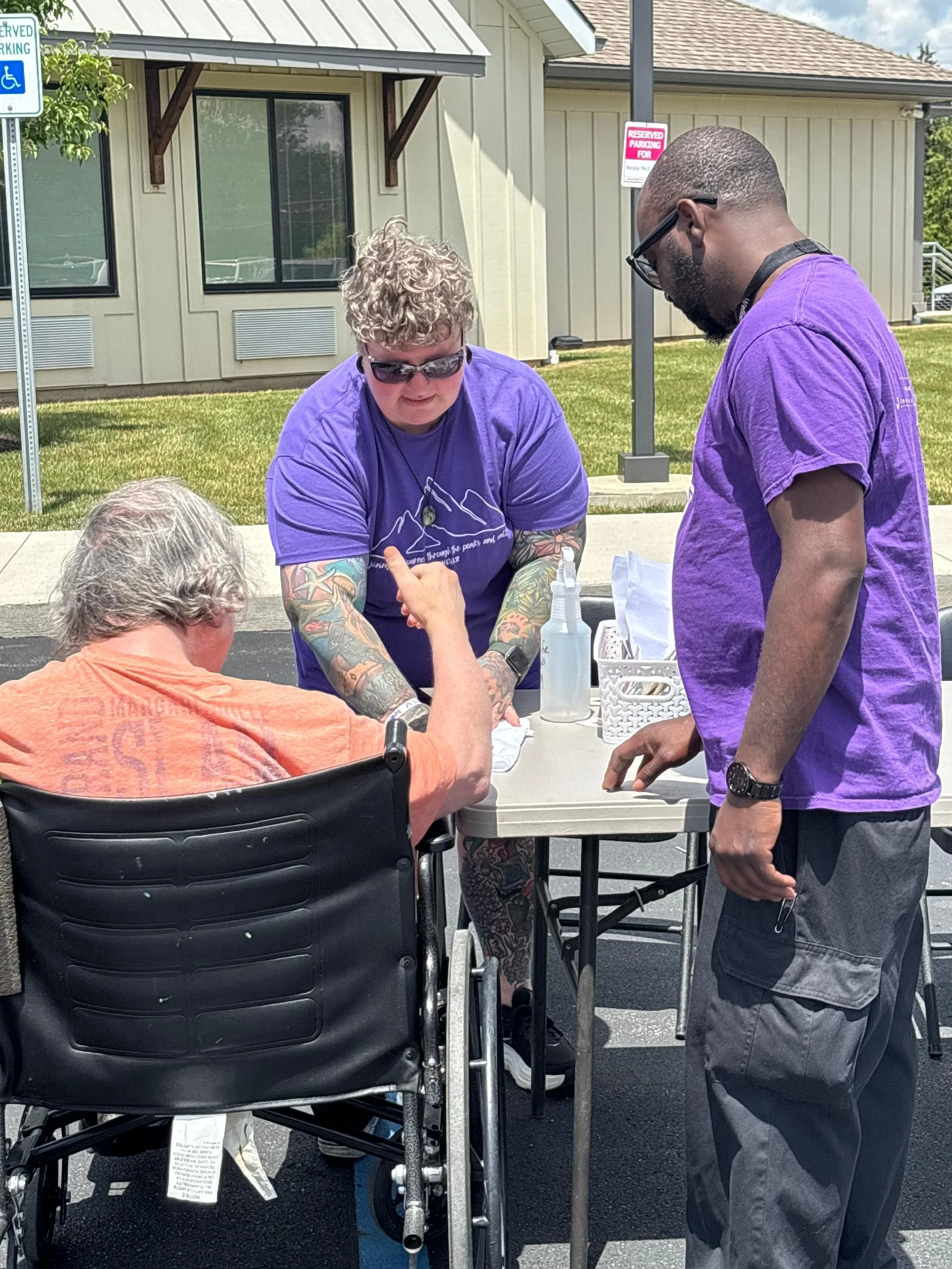 Two people, one with tattoos and sunglasses and another in a purple shirt, are talking to a person in a wheelchair at an outdoor table set up near a building on a sunny day.