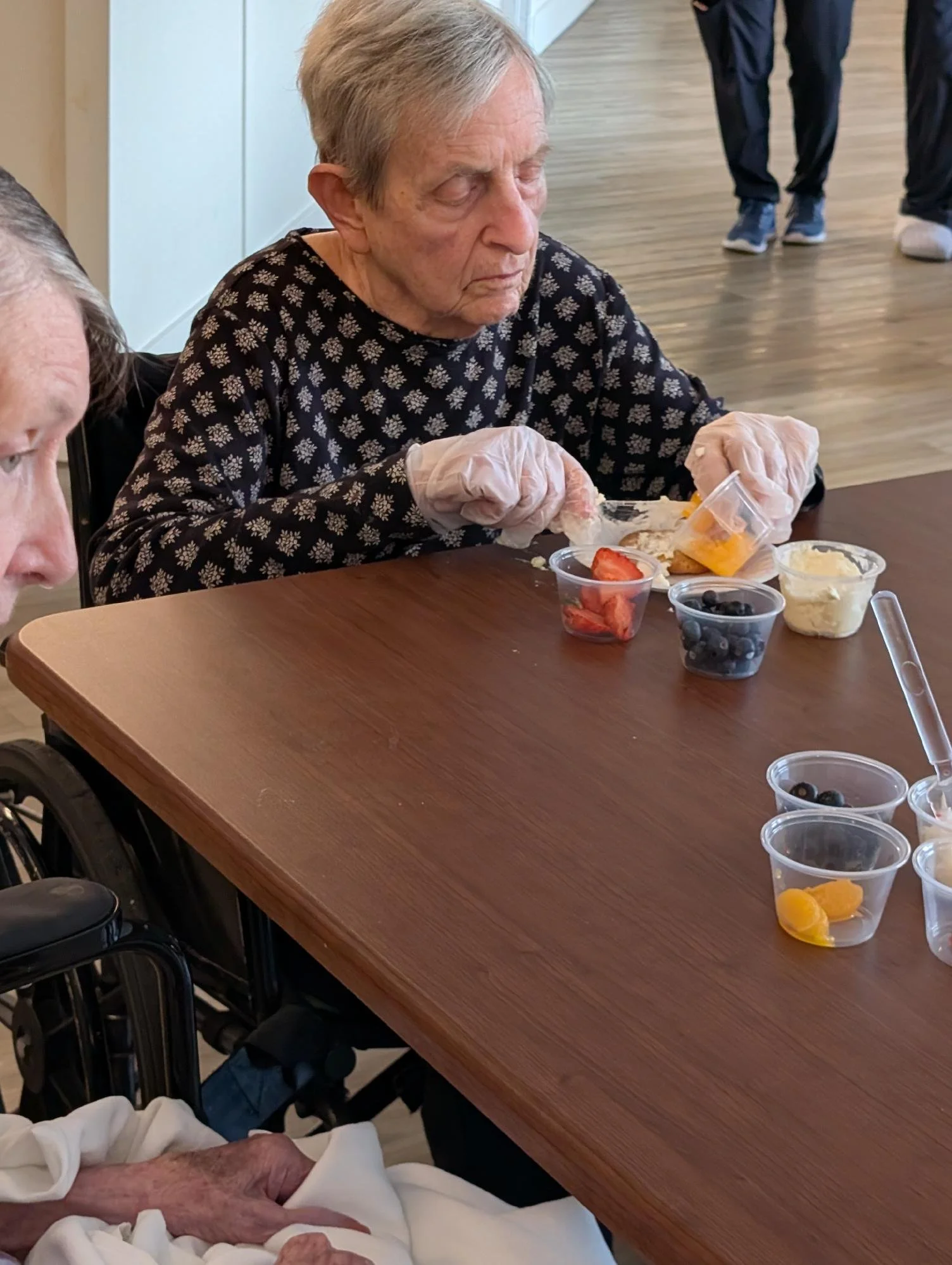 An elderly woman is sitting at a table, wearing a black sweater with a floral pattern, and is seen eating a meal with disposable gloves. There are small containers of food, including strawberries, blueberries, peach slices, and what appears to be mas