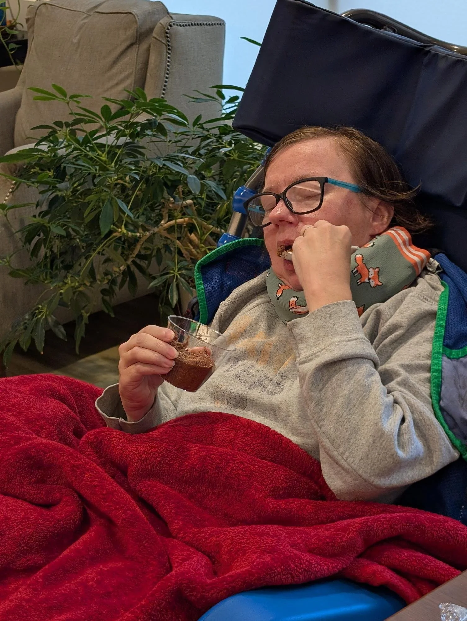 A person with glasses resting in a wheelchair, holding a drink, and wearing a bib with toy animals, covered with a red blanket, next to a green plant and a beige couch in a cozy room.