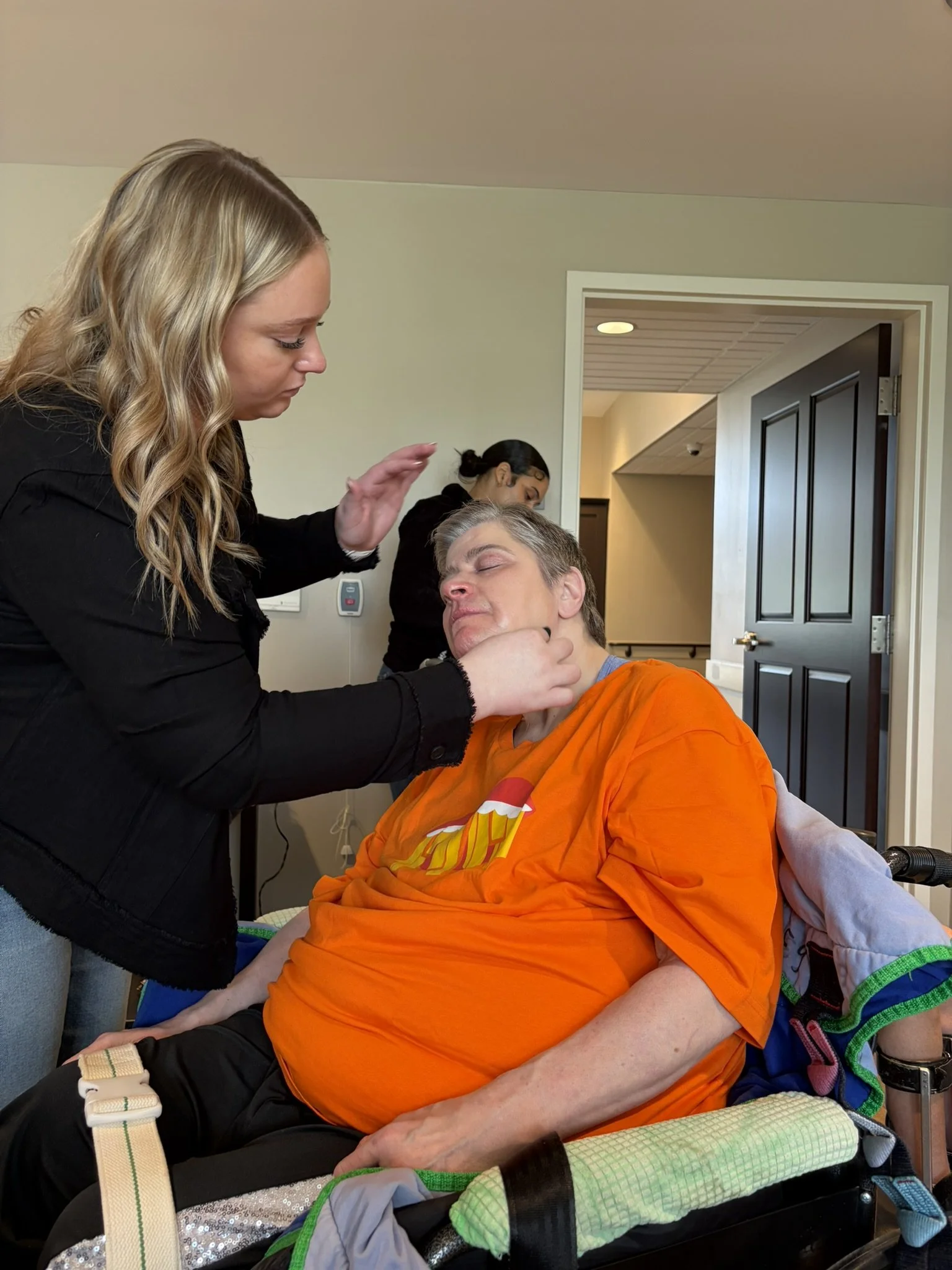 A woman with long wavy blonde hair giving a gentle stroke to an older man's face as he rests with eyes closed in a wheelchair, in a room with partially open doors and another woman in the background.