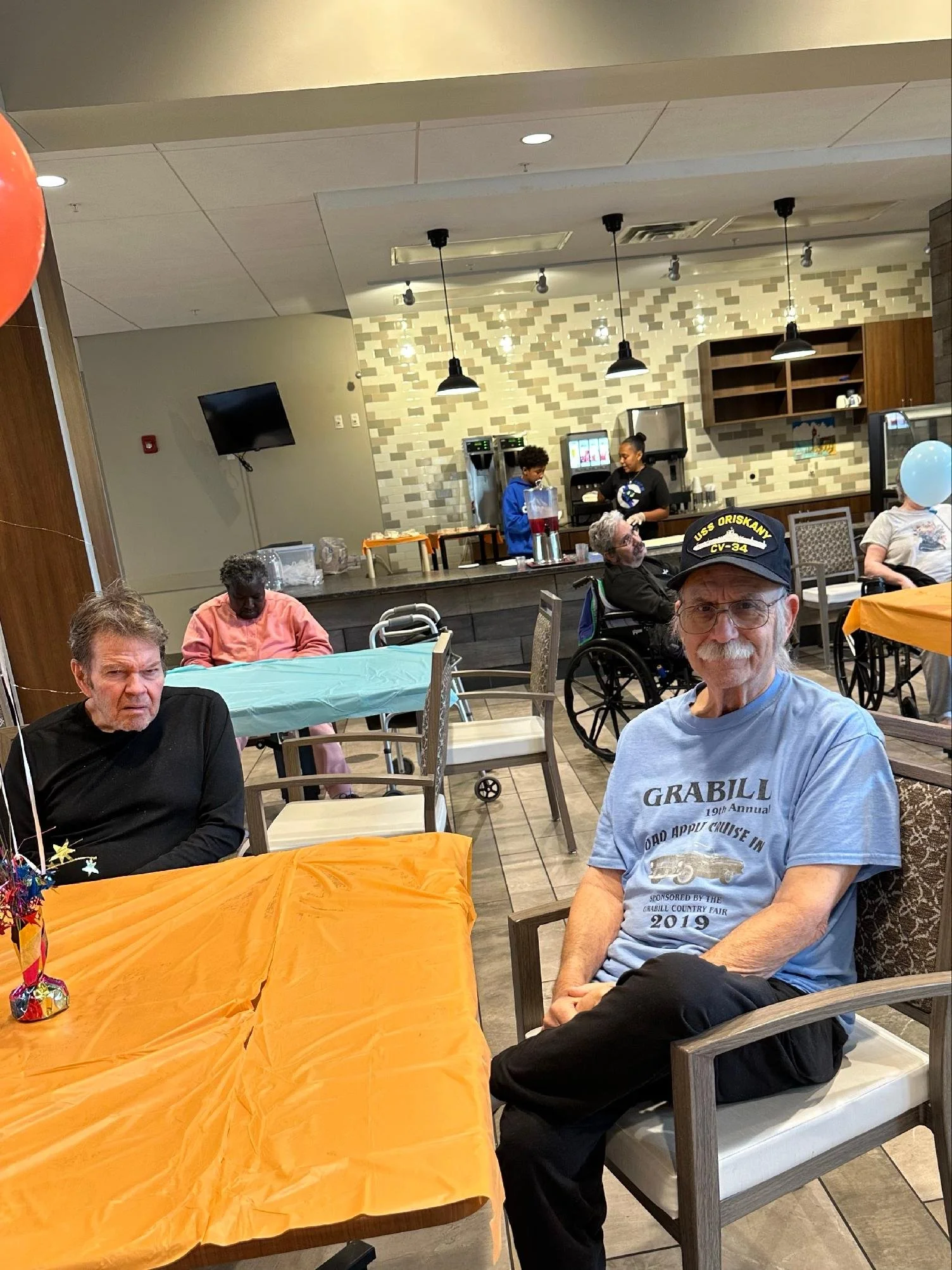 People gathered in a restaurant or event space, sitting around tables with orange and turquoise tablecloths, some in wheelchairs, with a dessert station in the background and decorative balloons on the side.