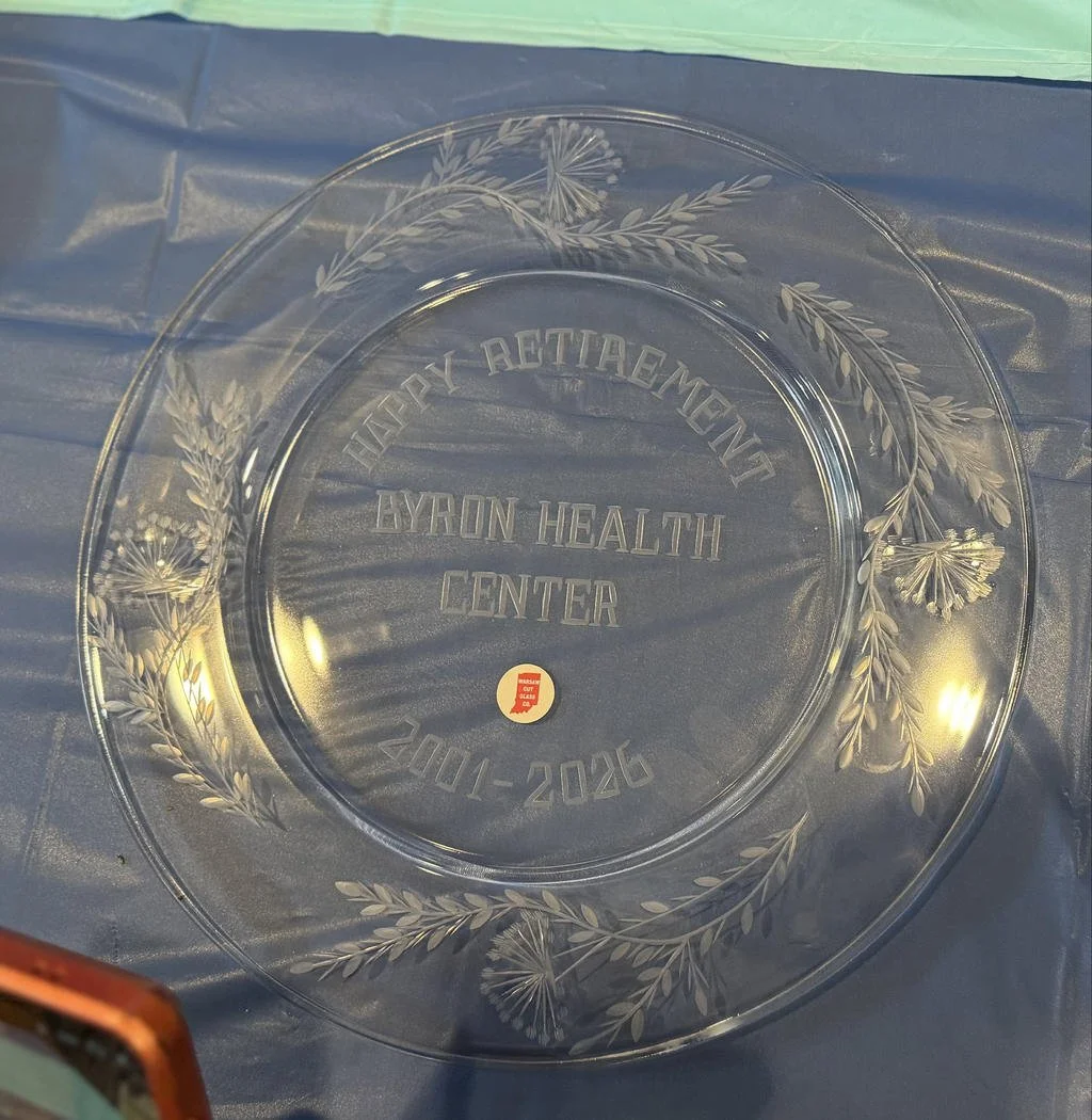 Clear glass plate with etched floral design and the inscription 'Happy Retirement, Byron Health Center, 2021-2026' in the center, placed on a blue surface.