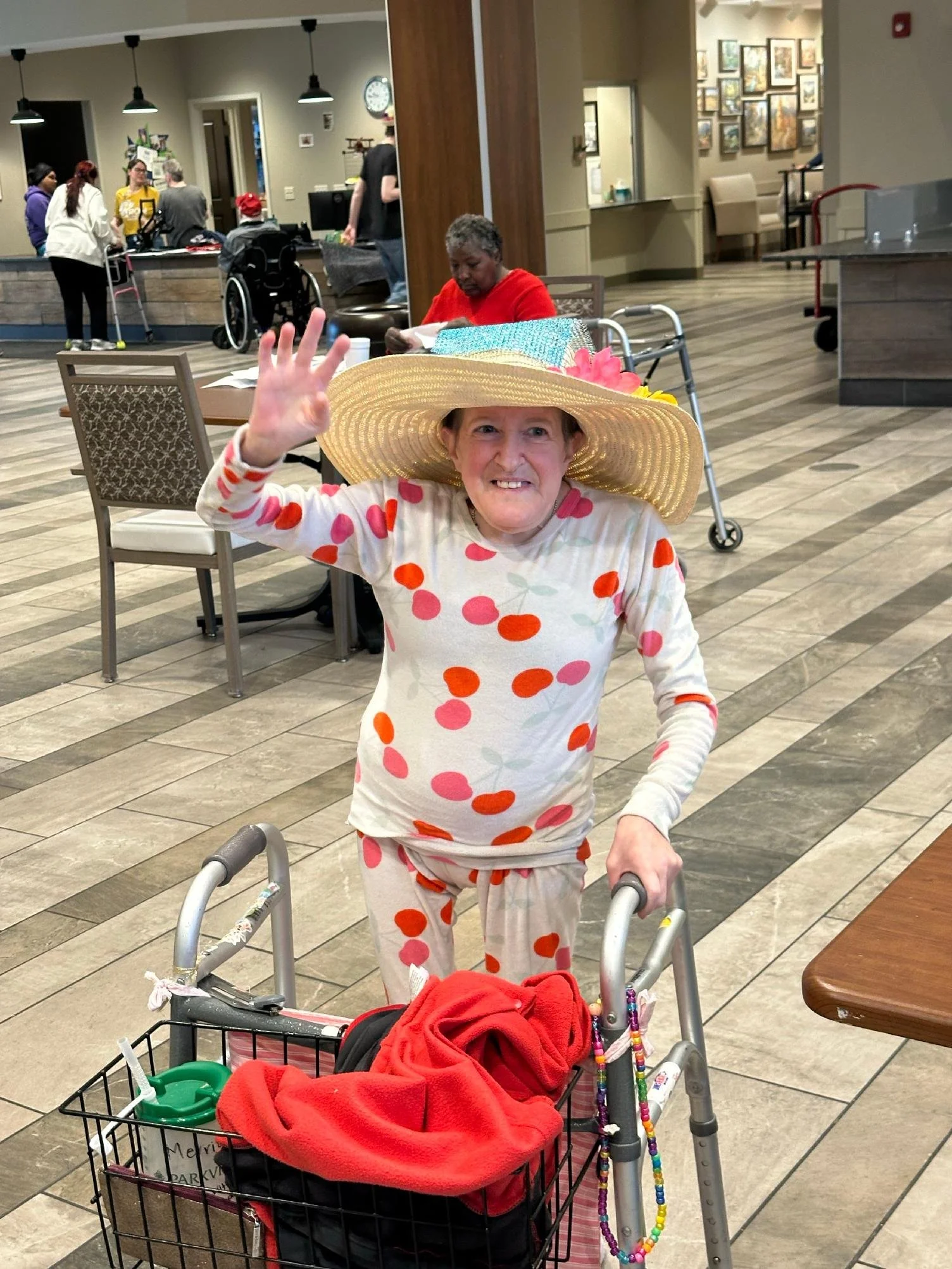 An elderly woman wearing a large straw sunhat with a flower and a bouquet of beads, waving and smiling at the camera. She is using a walker and surrounded by a shopping cart filled with clothing and blankets. In the background, another woman sitting 