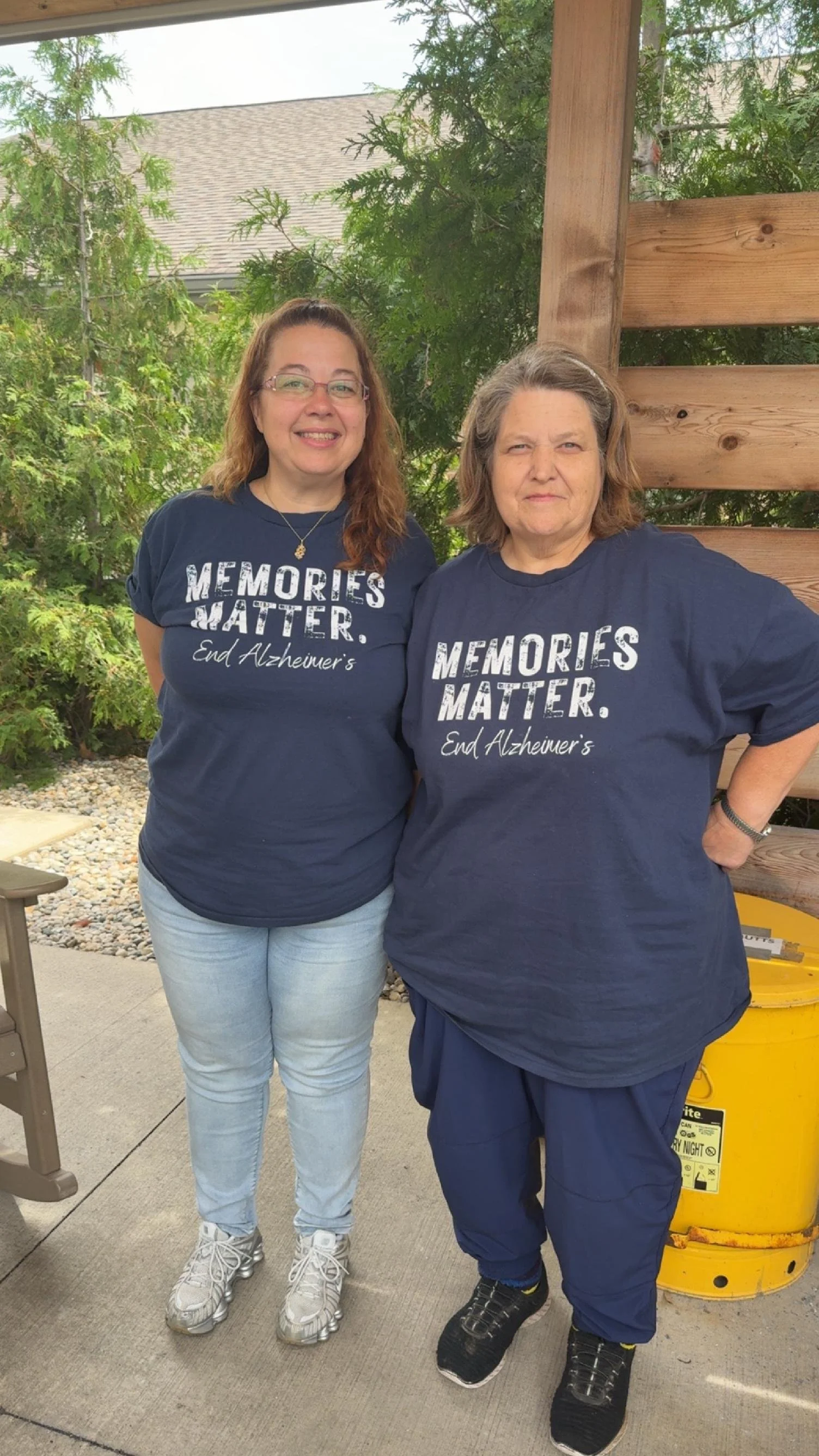 Two women wearing matching navy blue T-shirts with the text 'Memories Matter. End Alzheimer's' printed on them, standing outdoors next to a wooden fence and a yellow trash bin, smiling.