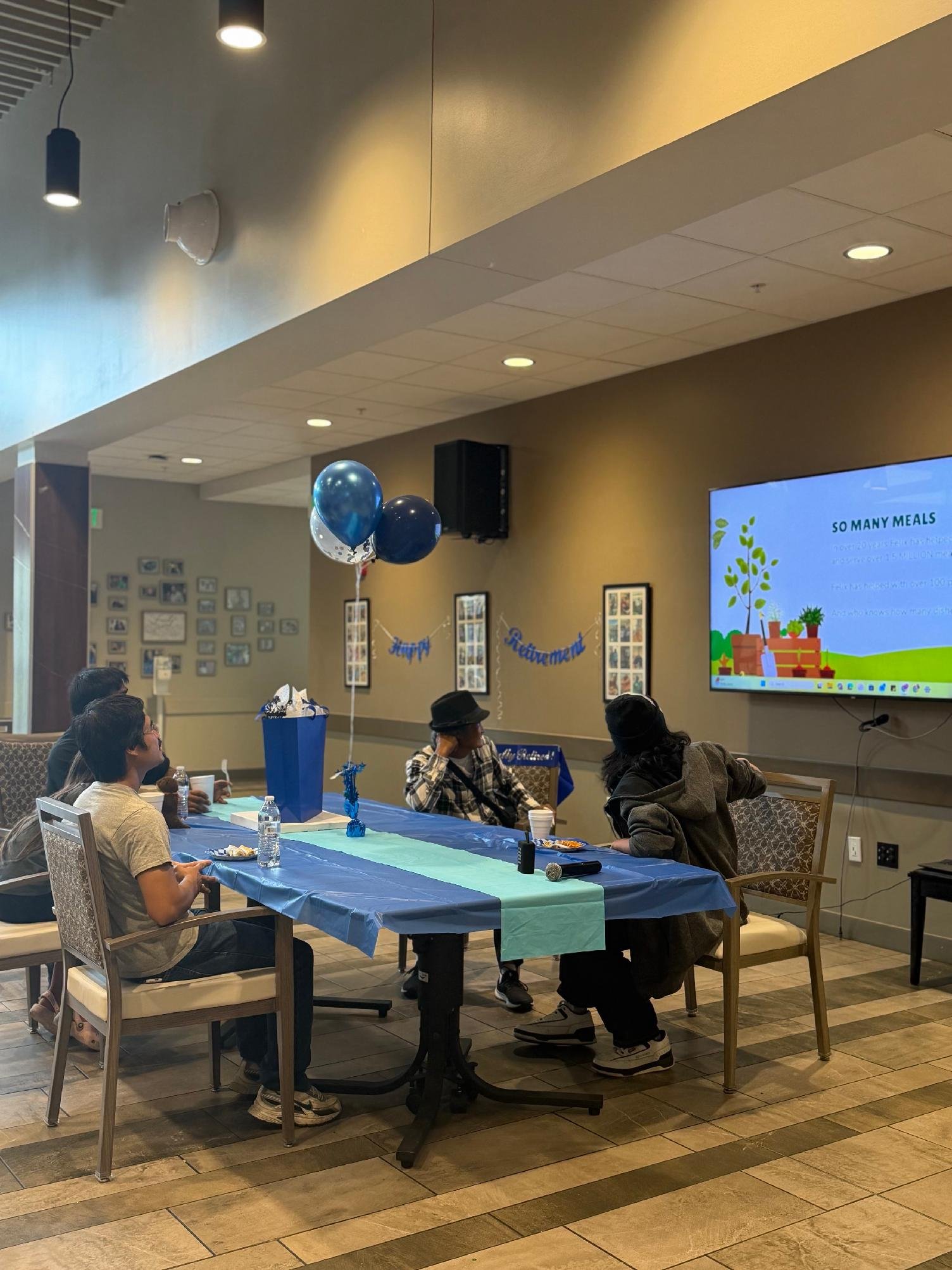 People sitting at a decorated table with balloons, watching a presentation on a large screen in an indoor event space.