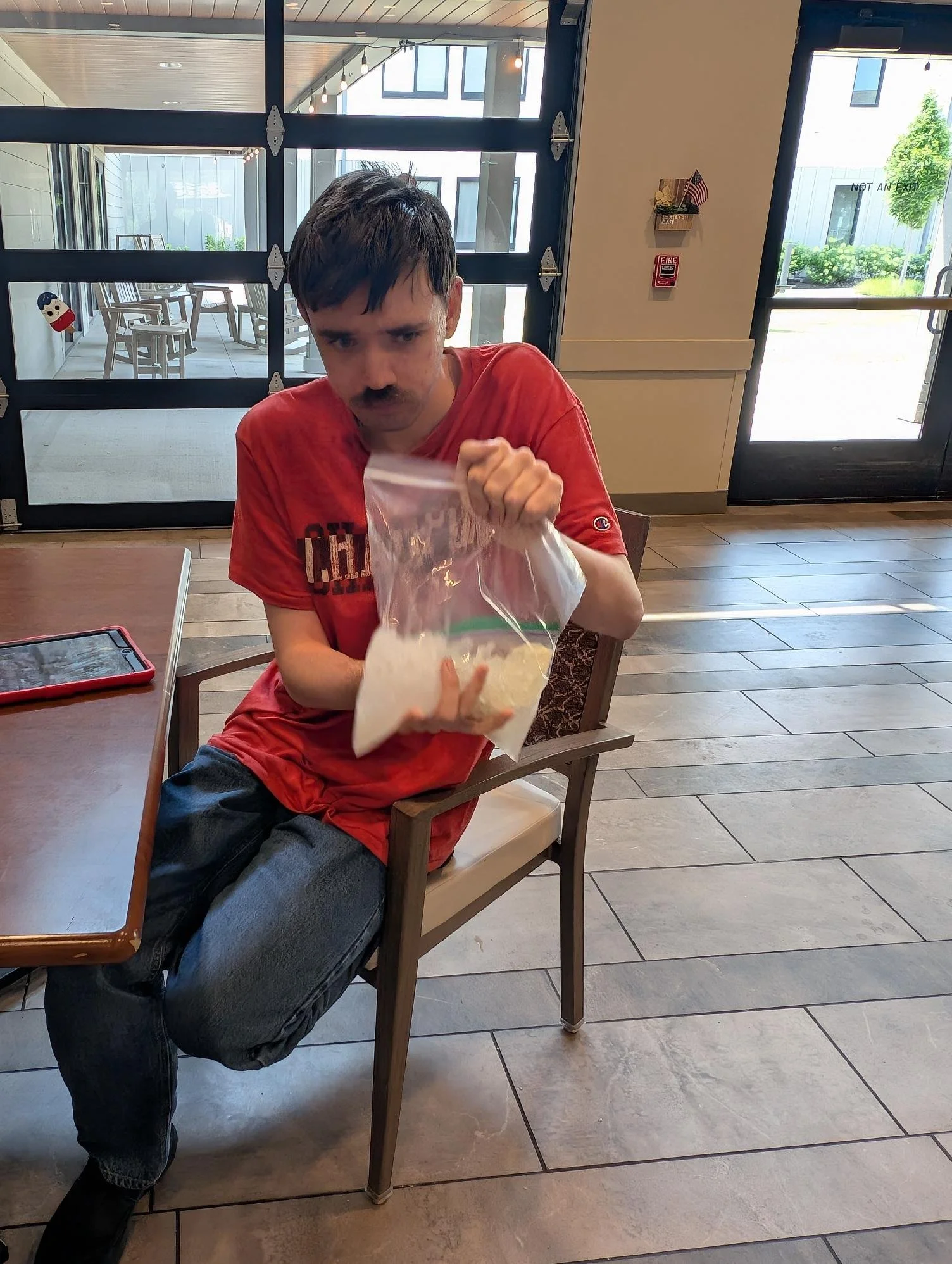 A man with dark hair, a mustache, wearing a red T-shirt and black pants, is sitting on a wooden chair in a restaurant or cafe. He is holding a plastic bag and looking directly at the camera with a serious expression. There is a tablet on the table in