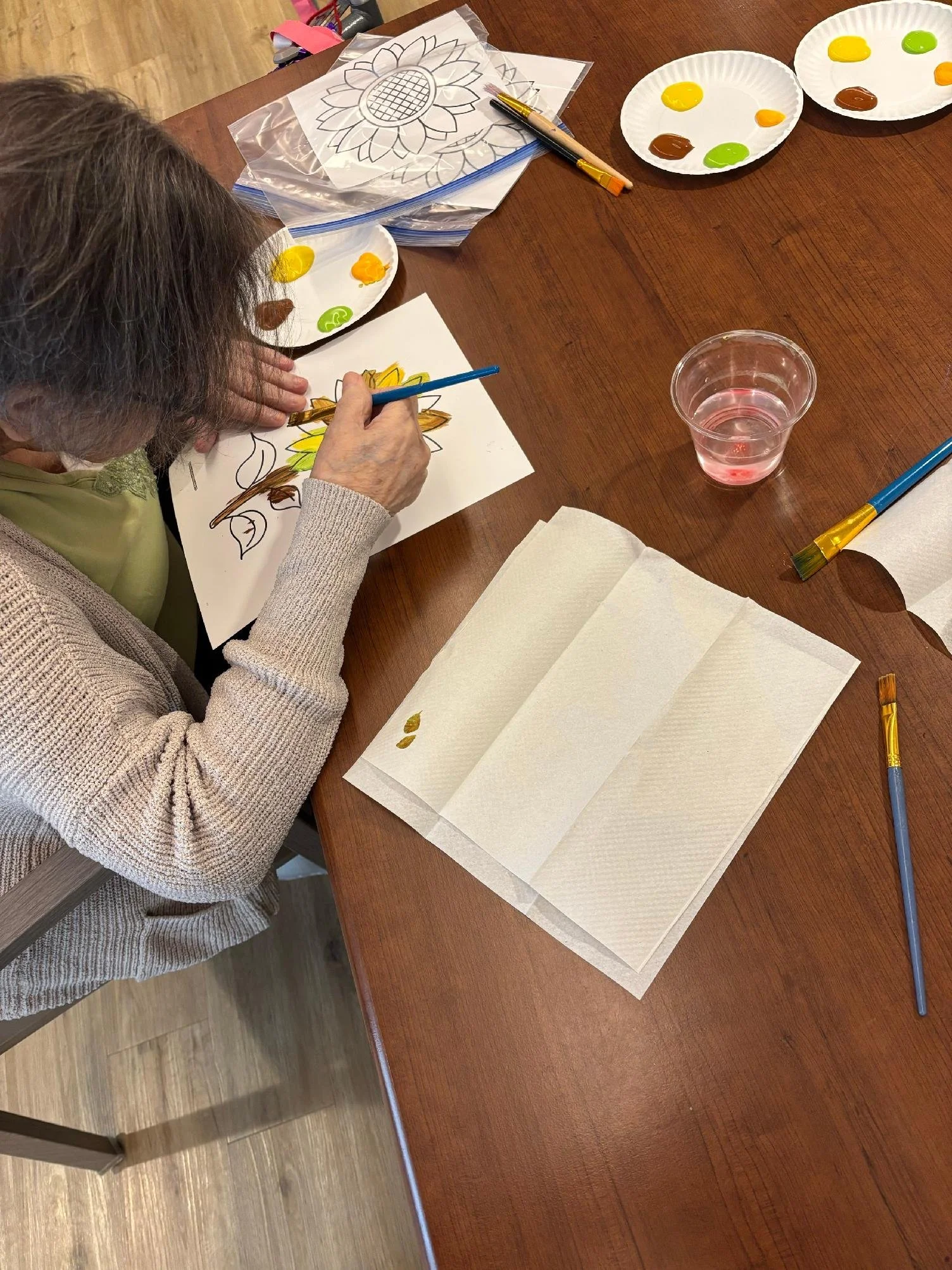 An elderly woman is sitting at a wooden table, painting a sunflower on paper using yellow, green, and brown paints. Several plates with paint spots, paintbrushes, and a plastic cup with water are on the table. There are also sheets of white paper and