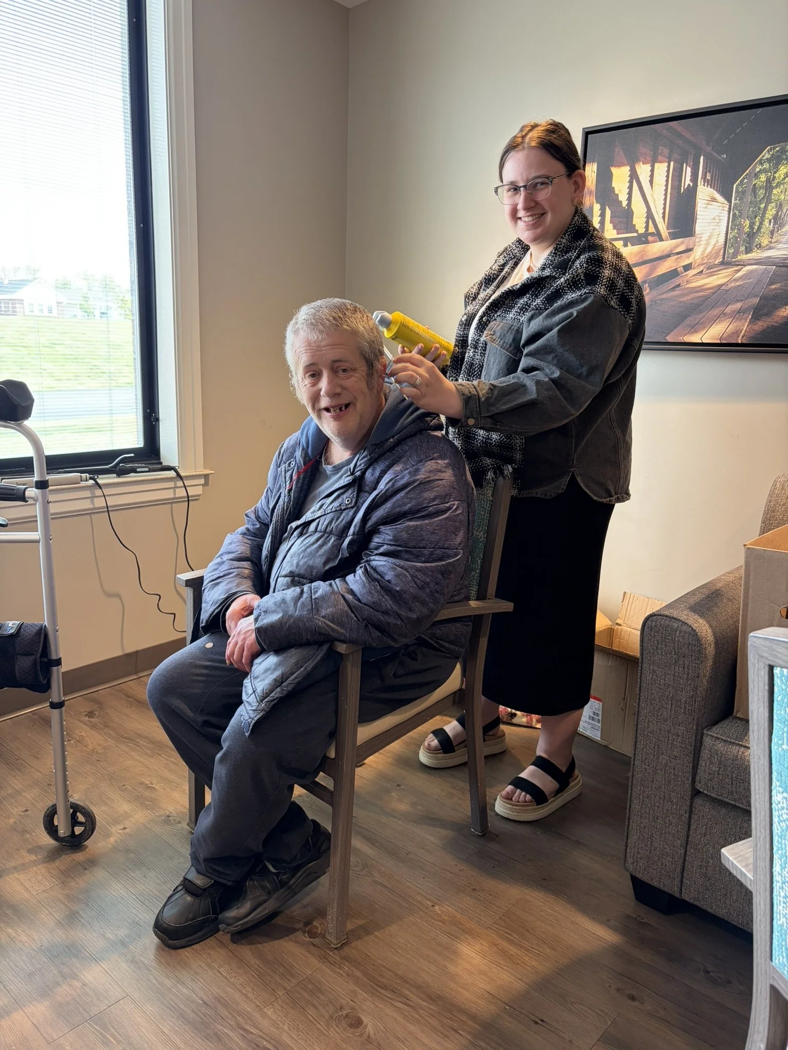 A woman with glasses is standing behind a seated man, holding a yellow spray bottle. The man has gray hair, is smiling, and has gaps between his teeth. They are in a room with wooden flooring, a window with blinds, and a framed picture of a wooden st