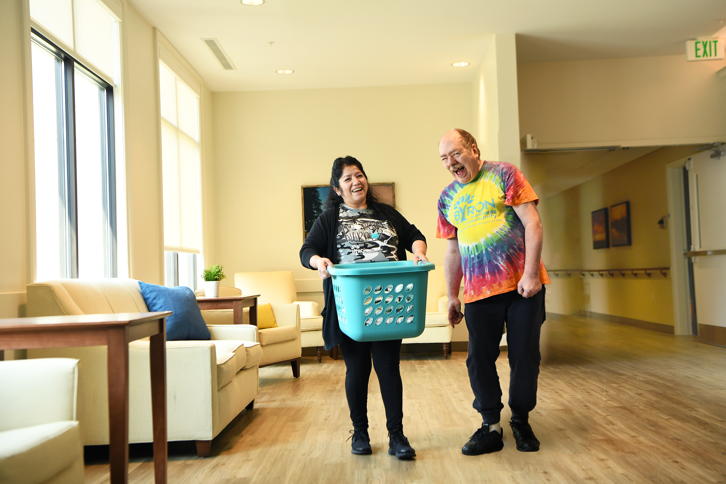 Two smiling people walking in a bright waiting room, one holding a laundry basket, the other wearing a colorful tie-dye shirt.