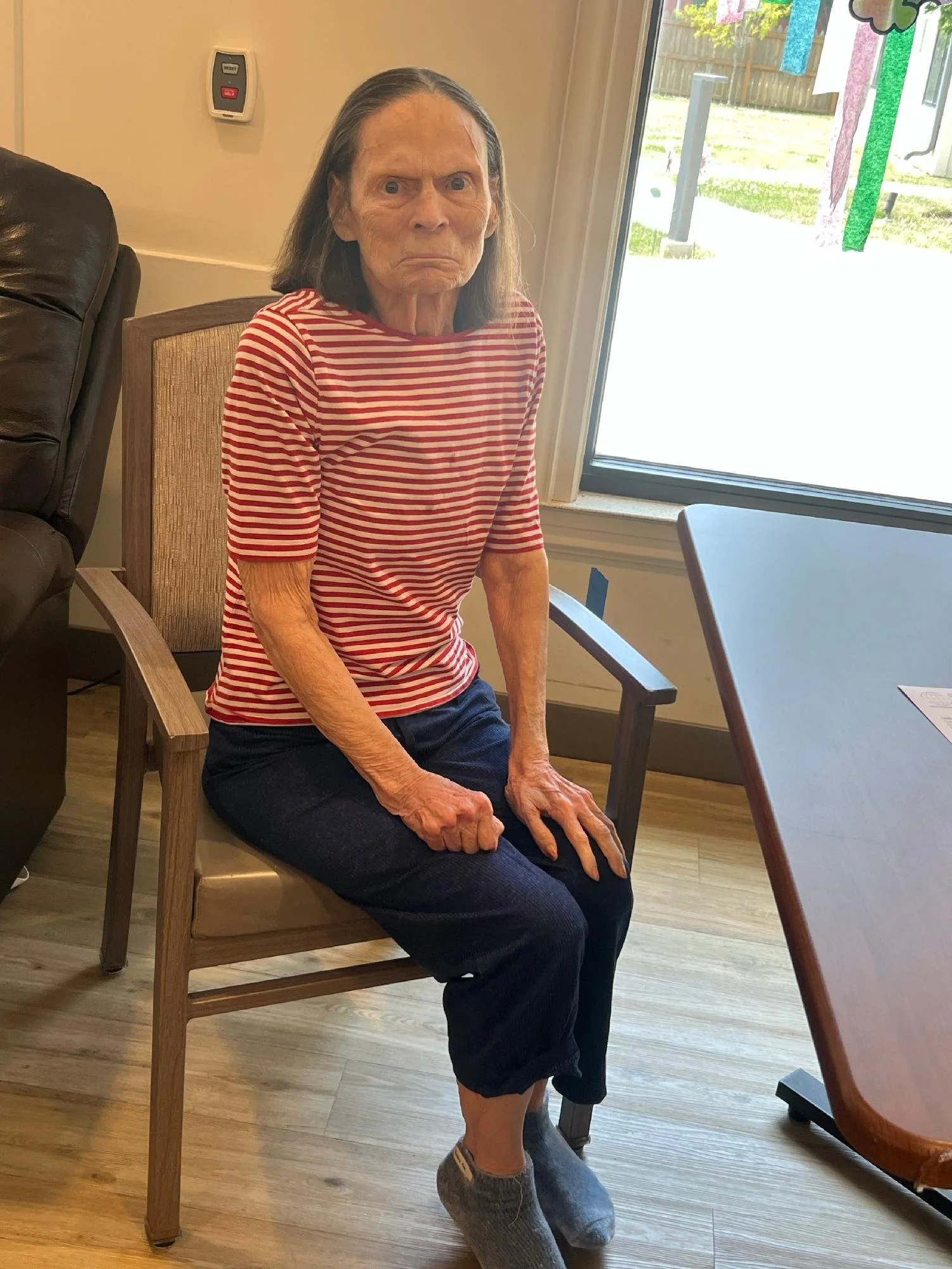 An elderly woman with wrinkled skin and long gray hair is sitting in a wooden chair with her hands resting on her knees. She is wearing a red and white striped shirt, dark pants, and slippers. She appears to have a serious or stern expression on her 