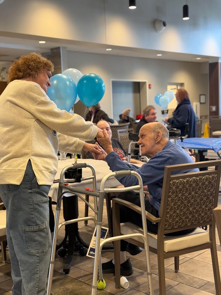 A woman with curly hair and beige sweater is holding hands and smiling at an elderly man with a walker, sitting at a table in a dining area decorated with blue balloons, while other people are in the background.