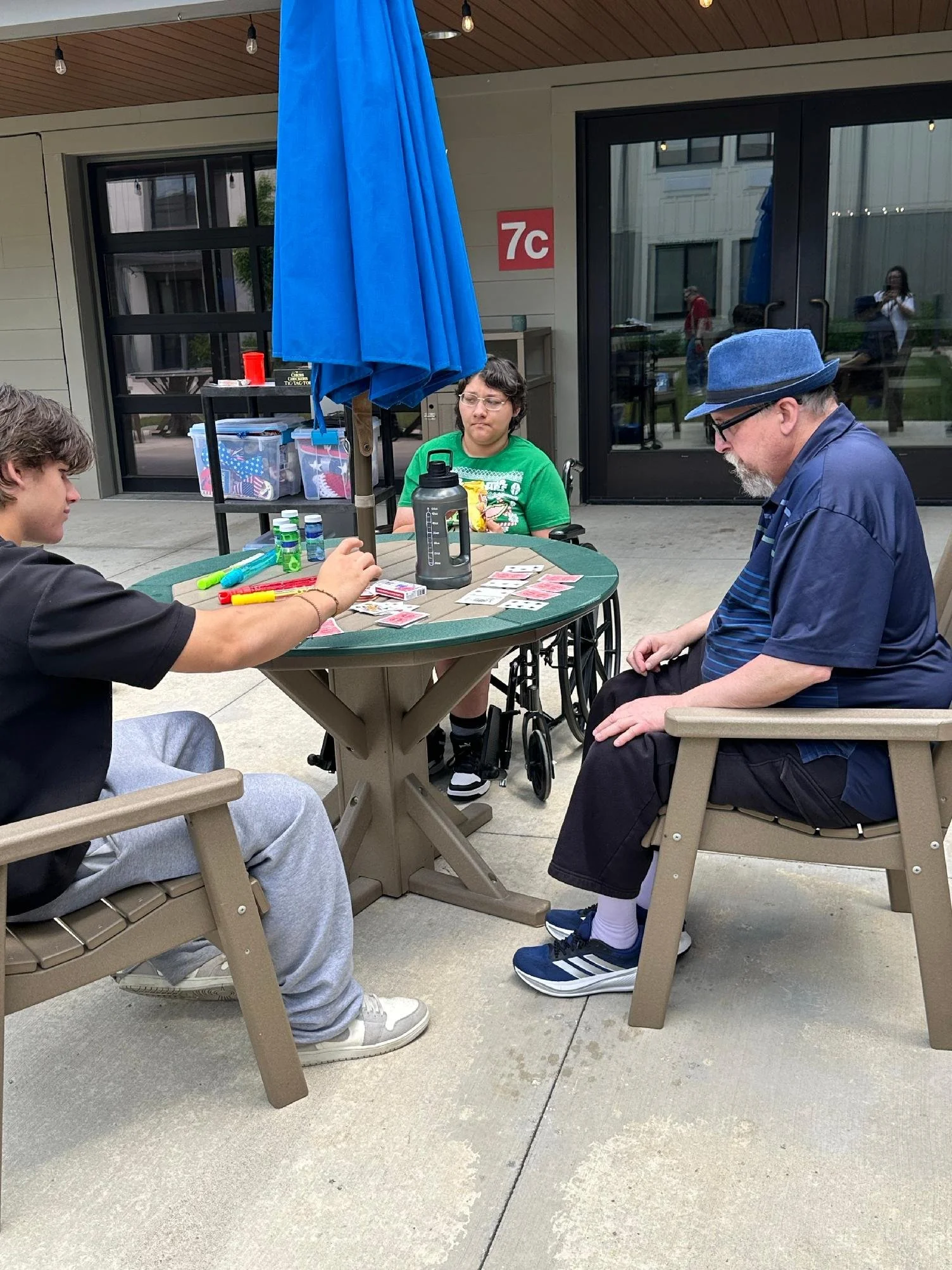 Three people sitting at a round table outside near a building with large glass doors. The person on the left is a young man in a black shirt with gray sweatpants, the person in the middle is a woman with glasses in a green shirt with a graphic design