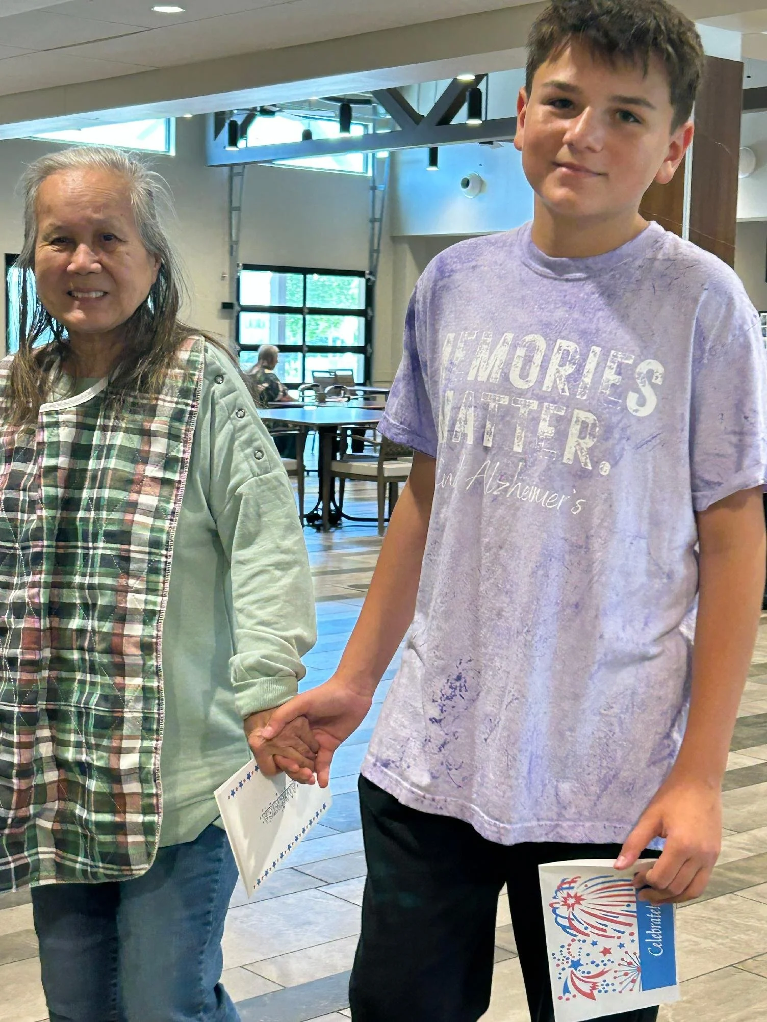 Two people holding hands; one appears to be an older woman and the other a young man. They are standing indoors, possibly in a public building or celebration venue, with tables and others visible in the background.