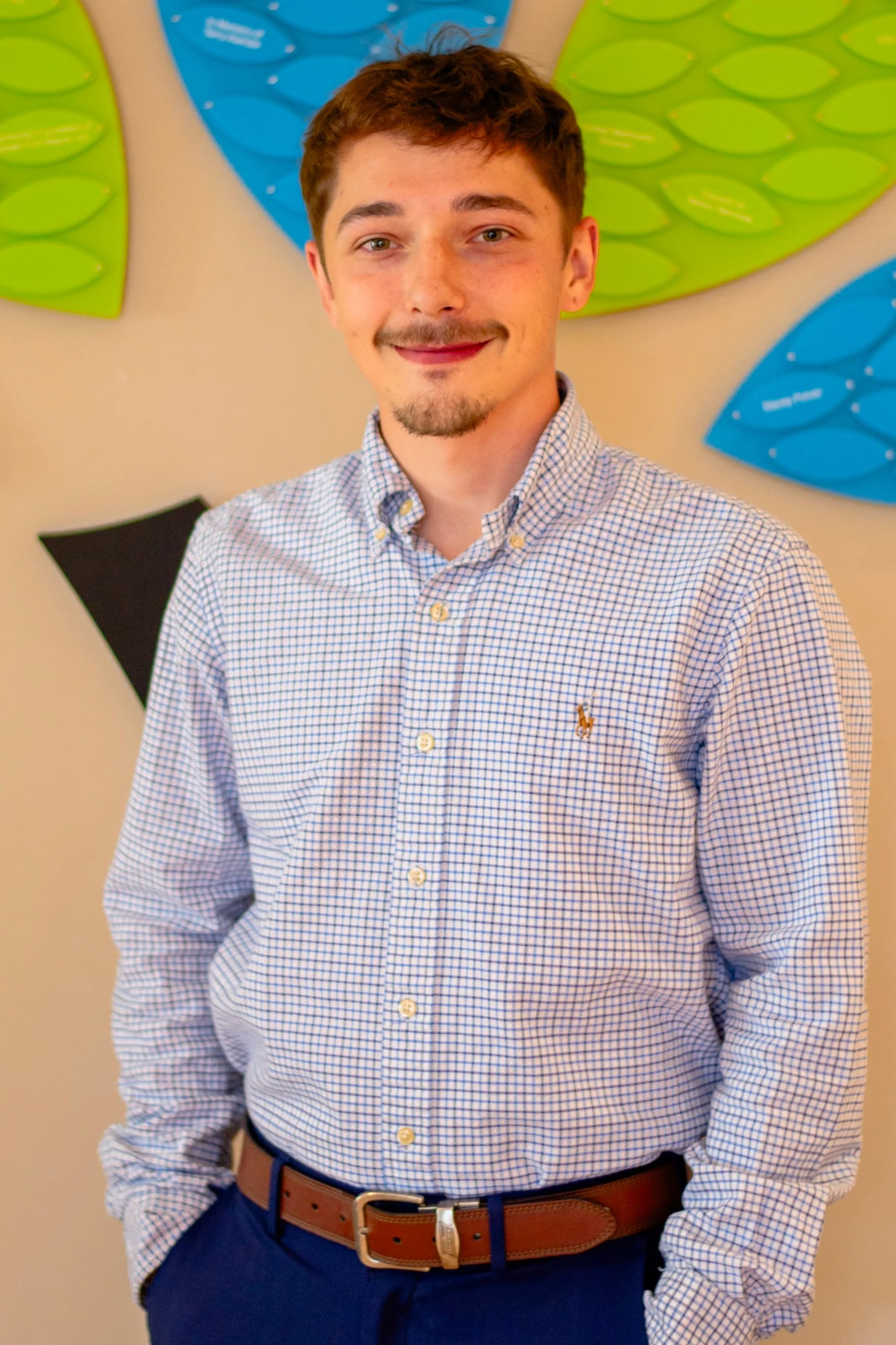 A young man with short brown hair, a goatee, and pale skin, smiling and dressed in a light blue checkered button-up shirt, gray belt, and navy pants. He is standing indoors against a wall with colorful abstract art featuring large green and blue shapes.